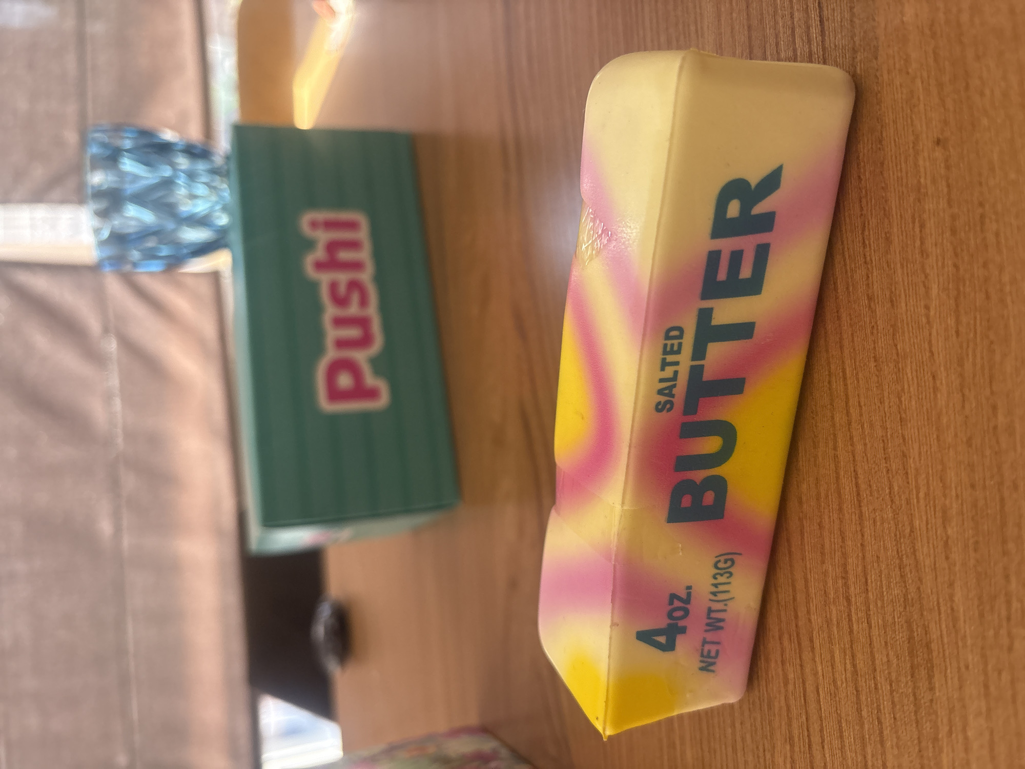 A close-up shot of a novelty butter-shaped item is positioned on a wooden table. The item features a swirled pattern of yellow, pink, and white, with the text "4 oz. SALTED BUTTER" and "NET WT.(113G)" printed in blue. In the background, a teal box with the word "Pushi" in pink lettering is visible, along with a blue faceted glass. The overall lighting suggests an indoor setting.