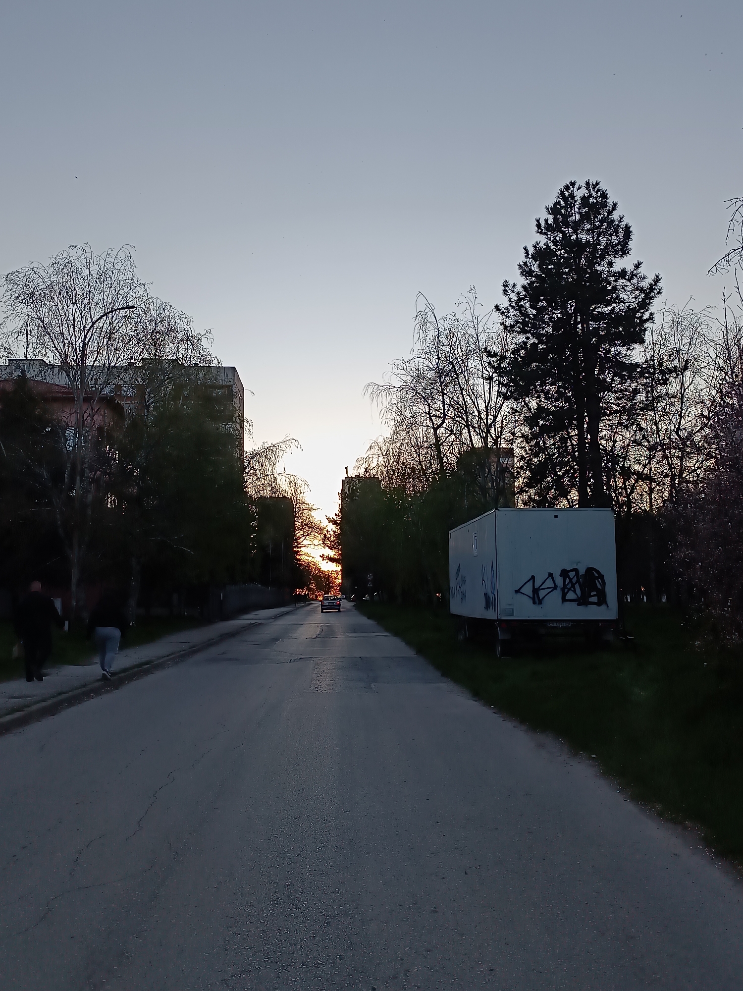 The image shows a road leading into the sunset in Pleven, Bulgaria. Buildings line the left side of the road, while a trailer with graffiti is on the right. Trees with bare branches are visible along both sides. Two people are walking on the left sidewalk. A car is driving towards the camera in the distance.