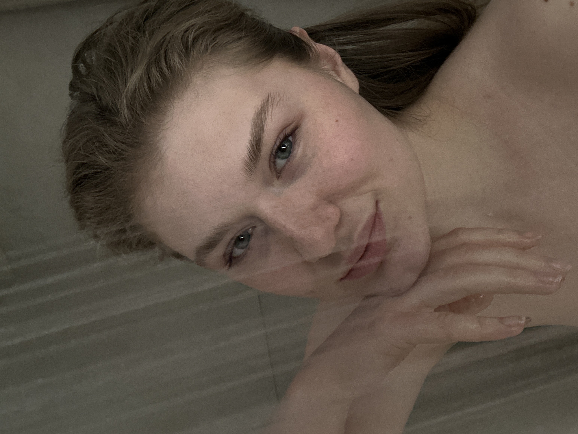 A close-up, chest-up portrait features a young adult female with fair skin, light brown hair, and light-colored eyes, appearing to be in a wet indoor environment, likely a shower. Her hair is pulled back and appears damp. Freckles are visible across her nose and cheeks. She looks directly at the camera with a slight, closed-mouth smile. Her right hand, visibly wet with water droplets, is bent at the wrist and rests with curled fingers against her jawline and neck. Her bare right shoulder is partially visible in the lower right of the frame, also appearing wet. The background is a uniformly colored, out-of-focus grey-green surface with faint vertical or diagonal lines, consistent with a tiled wall. The specific location within Kazan, Russia, cannot be determined from the visual information present in the image.