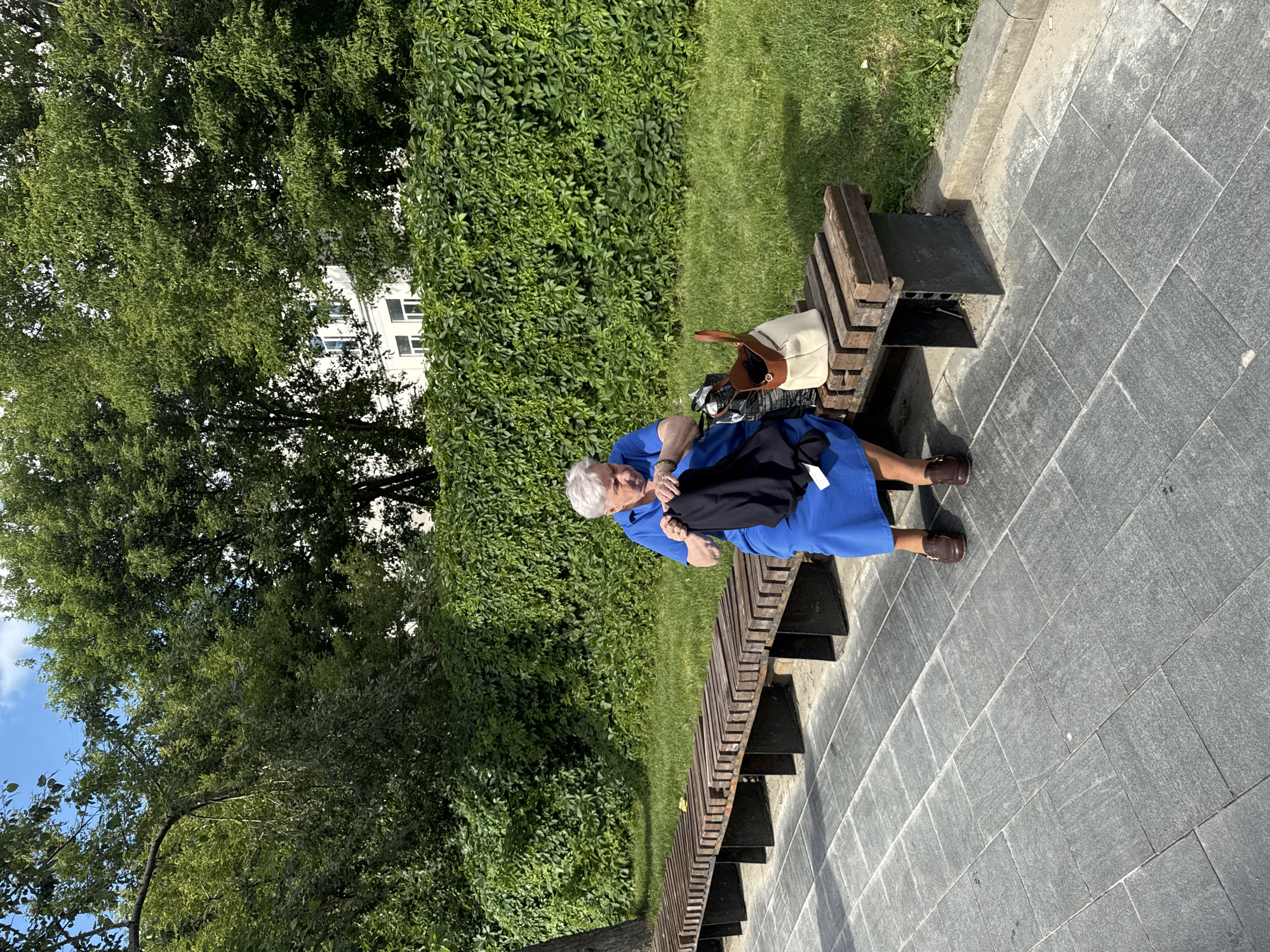 An elderly woman with short white hair is sitting on a wooden bench in a park. She is wearing a bright blue dress and brown shoes. In her hands, she holds a black garment, which she appears to be folding or adjusting. Next to her on the bench are two bags: a darker patterned bag and a lighter-colored leather bag with a brown handle. The bench is situated on a paved walkway, with a grassy area and a dense green hedge behind it. In the background, a white building with windows is visible, partially obscured by the leaves of trees. The sky above is blue with some clouds. The overall impression is a sunny day in a well-maintained urban green space.