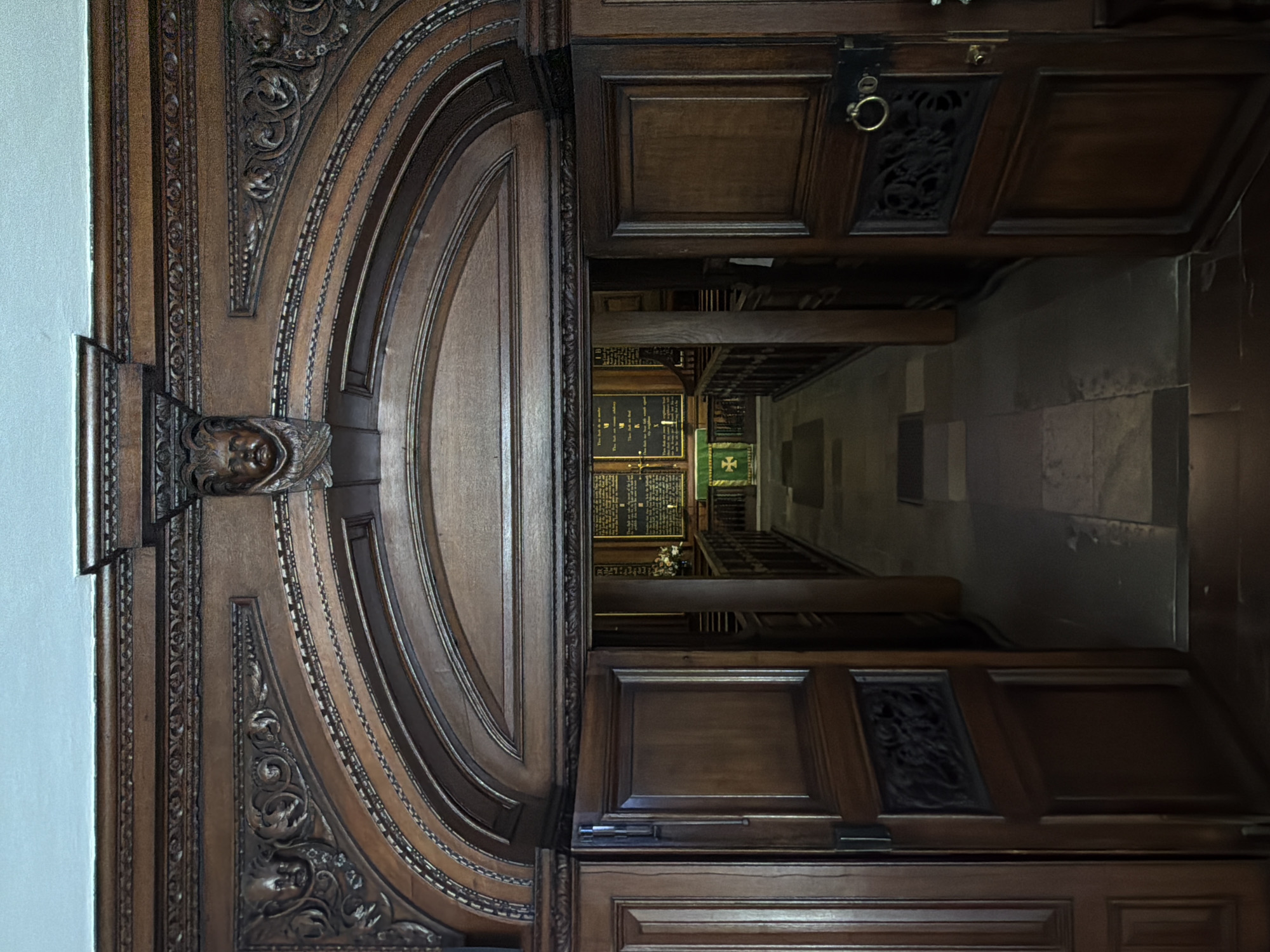 The image shows an ornate wooden doorway leading into a church interior. The doorway is elaborately carved with decorative moldings and features a carved cherubic face at the apex of the arch. The doors themselves are made of dark wood, with one door slightly ajar on the left and the other open on the right, revealing a view of the church's nave. Inside the church, pews are visible, and at the far end, an altar area is decorated with a green cloth and a white cross. The walls of the nave appear to be lined with framed texts or memorials. The floor inside the doorway is paved with large stone tiles. The overall scene suggests an entrance to a historic church, likely within London, United Kingdom, given the architectural style and context.