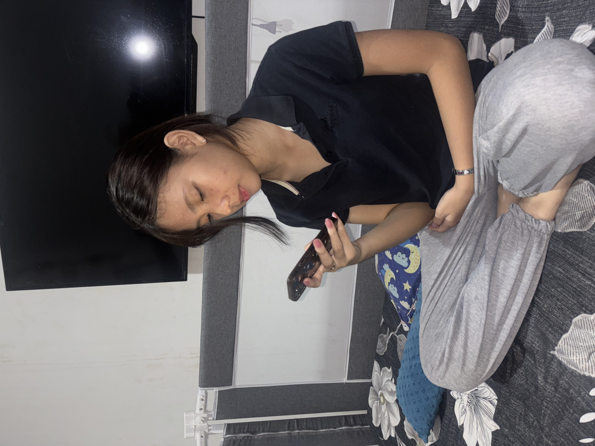 A young Asian female is sitting cross-legged on a bed, holding a smartphone in her right hand and looking down at it. She is wearing a black collared t-shirt and gray sweatpants. The bed has floral-patterned sheets and pillows. In the background, a large flat-screen television is mounted on the wall above the bed's headboard. The room appears to be a bedroom. The image was taken in West Jakarta, Indonesia.