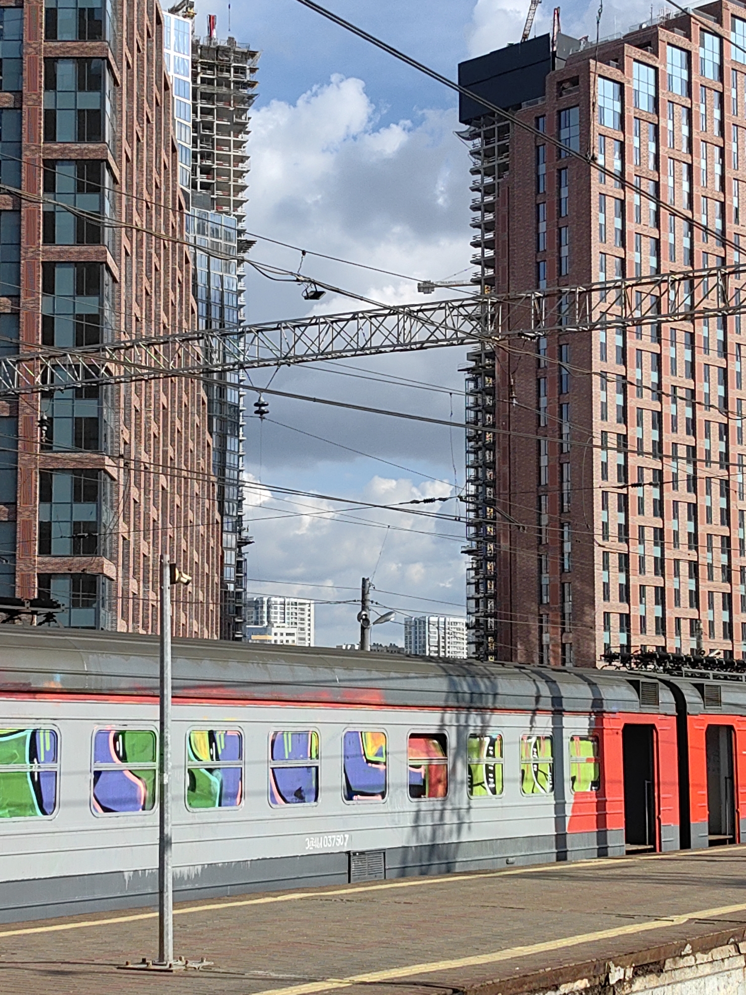 The image depicts a stationary train at a platform in Moscow, Russia, against a backdrop of modern buildings, some still under construction. The train cars are painted in gray and red, with graffiti reflected in the windows. Overhead power lines run above the train. The buildings in the background are high-rise structures, primarily brick, with visible construction scaffolding on some. The sky is partly cloudy. The platform is made of stone or concrete, showing signs of wear and tear.