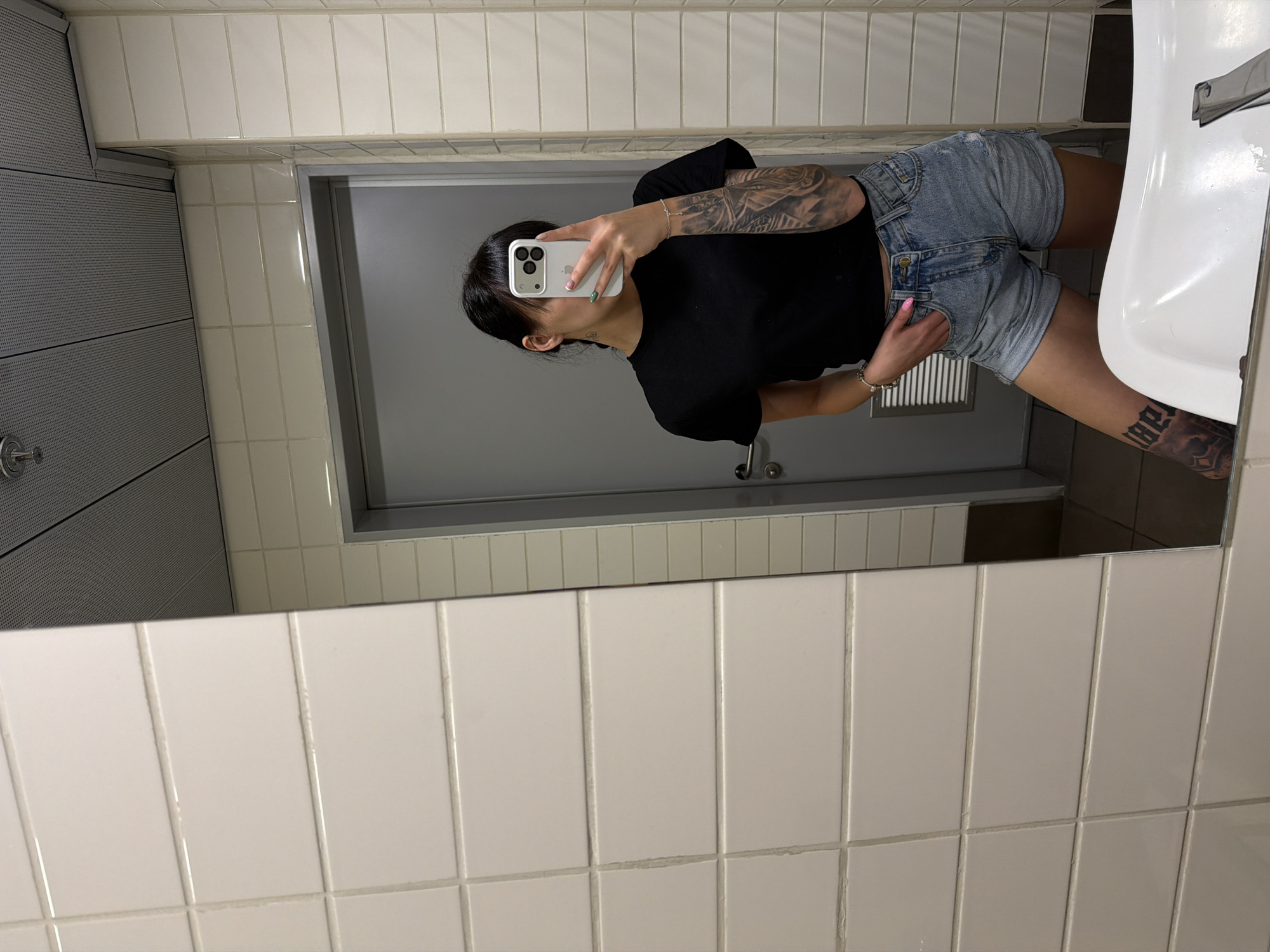 A mirror selfie captures an individual in what appears to be a public restroom in Plovdiv, Bulgaria. The person, seen from the chest down, wears a black t-shirt and light blue distressed denim shorts. They have dark hair pulled back and are holding a white smartphone with multiple cameras to take the picture, obscuring their face. Their left arm features a prominent sleeve tattoo with intricate designs, while a block lettering tattoo is visible on their right leg, just above the knee, though the specific text is not clearly legible. A silver bracelet adorns their left wrist, and a ring is visible on their right hand, which is casually placed in the front pocket of their shorts. The fingernails on the visible hand show light pink and multi-colored polish.
The background, reflected in the large mirror, reveals standard restroom features. The walls are predominantly covered with light-colored rectangular tiles, arranged vertically, with some sections having smaller grey rectangular tiles above them. A grey door is visible in the reflection, possibly a stall door or the entrance. To the right of the person, a white ceramic sink is partially visible. On the left side of the reflection (corresponding to the right side of the actual room), grey perforated panels suggest a ventilation system or ceiling element. The indoor lighting indicates an indeterminate time of day. The scene is static, focused on the posed selfie.