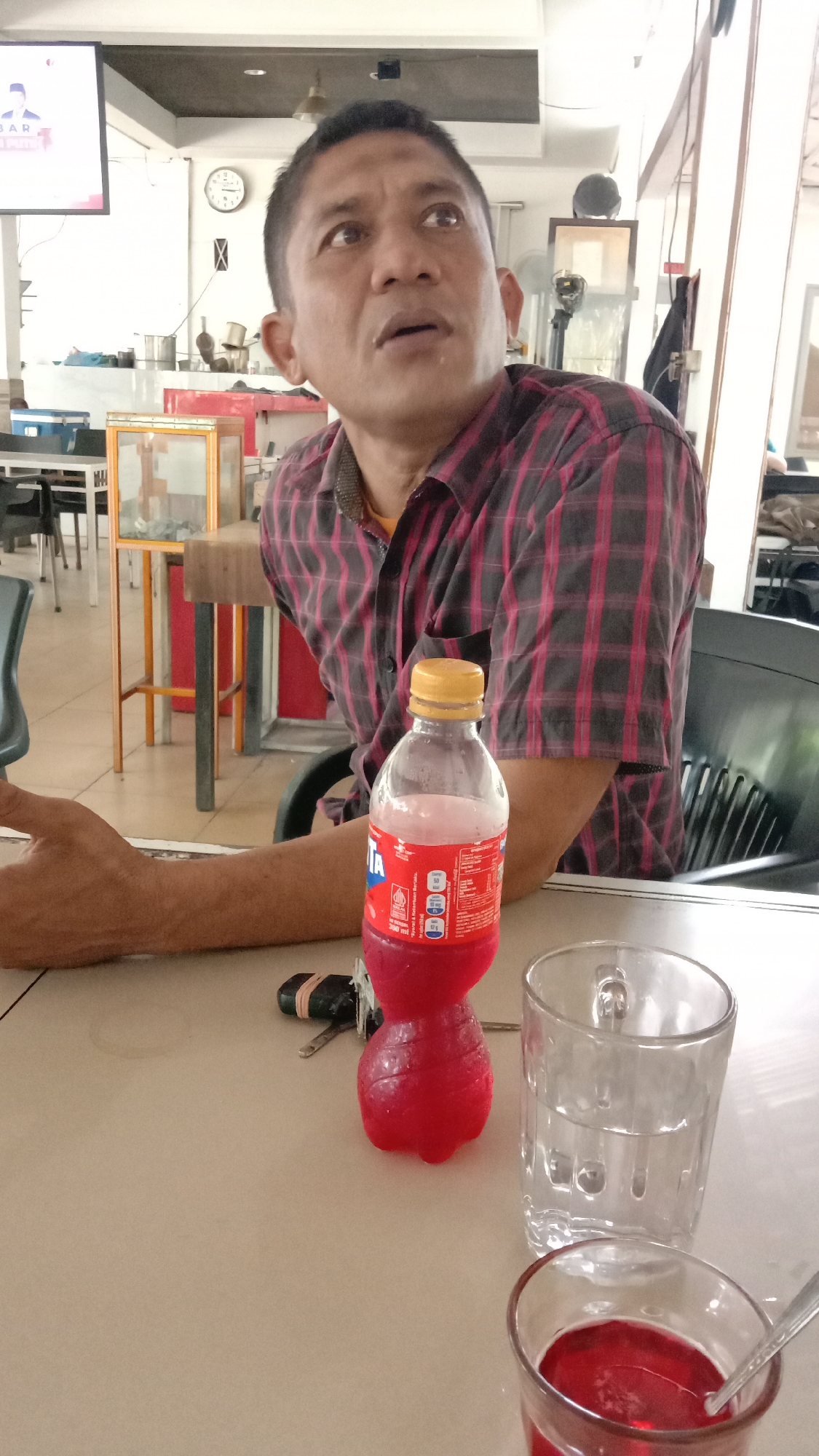 This image captures an indoor scene, likely in an eatery or cafe in Lhokseumawe, Indonesia, during the daytime. The primary subject is a man of medium build, seen from the chest up, seated at a table. He has short, dark hair and is wearing a red and black plaid button-up shirt over a yellow undershirt. His head is turned slightly to the right, and his gaze is directed upwards and to the right, with his mouth slightly open, suggesting he is observing or listening intently to something off-camera.
On the light-colored table in front of him, a small, red plastic bottle of Fanta, partially filled with red liquid, is prominently displayed. The bottle's label is partly visible, showing the brand name "FANTA" and Indonesian text indicating nutrition information ("PORSI & INFORMASI GIZI"). Next to the bottle are a set of keys. Two glasses are also on the table: a clear glass contains water, and a smaller glass holds a red liquid with a spoon in it, suggesting a beverage. The man's right arm and hand are resting on the table.
The background reveals a brightly lit interior space with white walls. Various elements typical of a public eating establishment are visible, including tables, chairs, and a display cabinet. A wall-mounted clock indicates the time is approximately 1:30. In the upper left, a television screen displays what appears to be a news or political broadcast, featuring a person's image and partially visible text, including the word "BAR". The scene conveys a casual, everyday atmosphere.