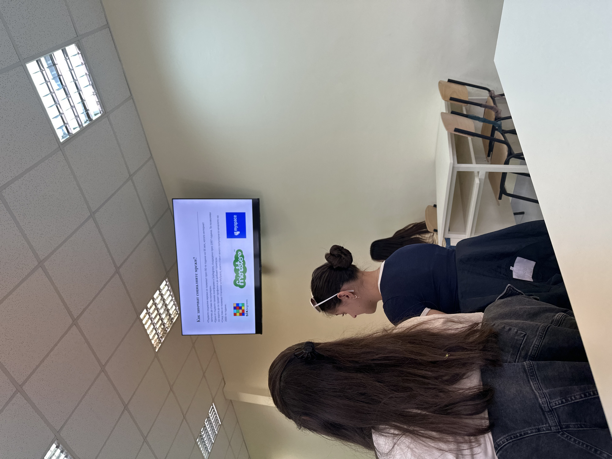 The image shows an indoor classroom setting, likely in Sofia, Bulgaria, given the Bulgarian text on the TV screen. Two women are in the foreground, presumably students. One has long dark hair and is wearing a white shirt under a dark denim jacket. The other woman has her hair in a bun, wears white framed sunglasses, and is in a navy blue shirt. Both appear to be seated at desks. A row of empty chairs is visible behind the women, stacked neatly along a wall with a white table. A TV screen is mounted on the wall, displaying information about the history of social networks. The ceiling is a grid pattern with recessed fluorescent lighting fixtures.