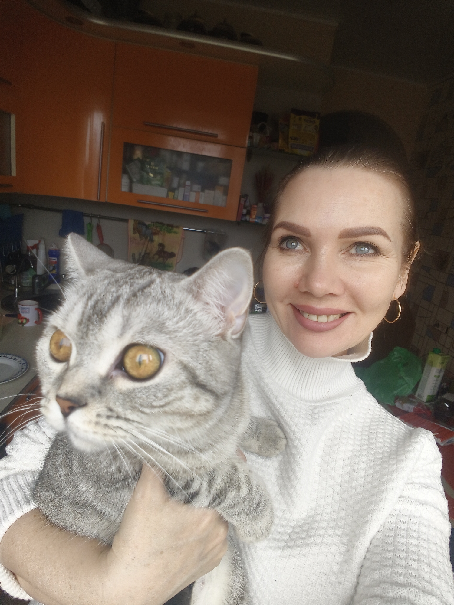 The image is a close-up selfie of a smiling woman holding a cat in an indoor setting, likely a kitchen. The woman has light blue eyes, fair skin, and light brown hair pulled back. She is smiling broadly, showing a pleasant expression, and is wearing a white, textured turtleneck sweater and gold hoop earrings.
She is holding a grey tabby cat with prominent darker stripes and large, striking amber-yellow eyes. The cat is looking intently towards the left side of the frame, away from the camera, and appears calm in her arms.
The background shows a domestic interior with bright orange kitchen cabinets, some featuring silver handles. One cabinet has a glass front, revealing various items, including bottles and boxes. Below the cabinets, parts of a kitchen counter and a sink area are visible, along with a faucet, a dish rack, a white mug with a red design, and a patterned hand towel. To the right, a green plastic bag and a white carton (possibly a drink container) are partially visible. The lighting suggests it is daytime. No distinct, legible text is visible on any items in the background.