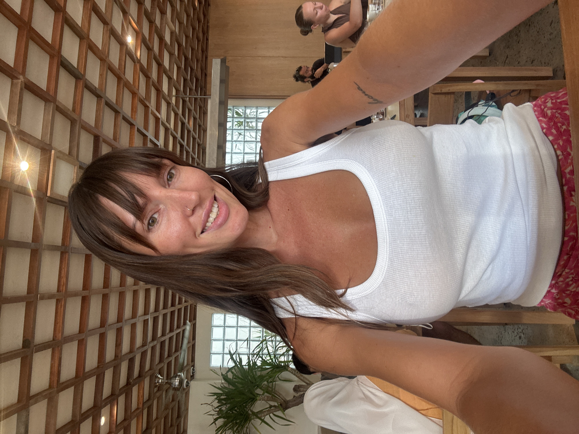A woman is taking a selfie in a restaurant in North Kuta, Indonesia. She has brown hair and bangs, and is wearing a white tank top. The restaurant has a wooden lattice wall and a green plant in the background. There are other people in the restaurant.