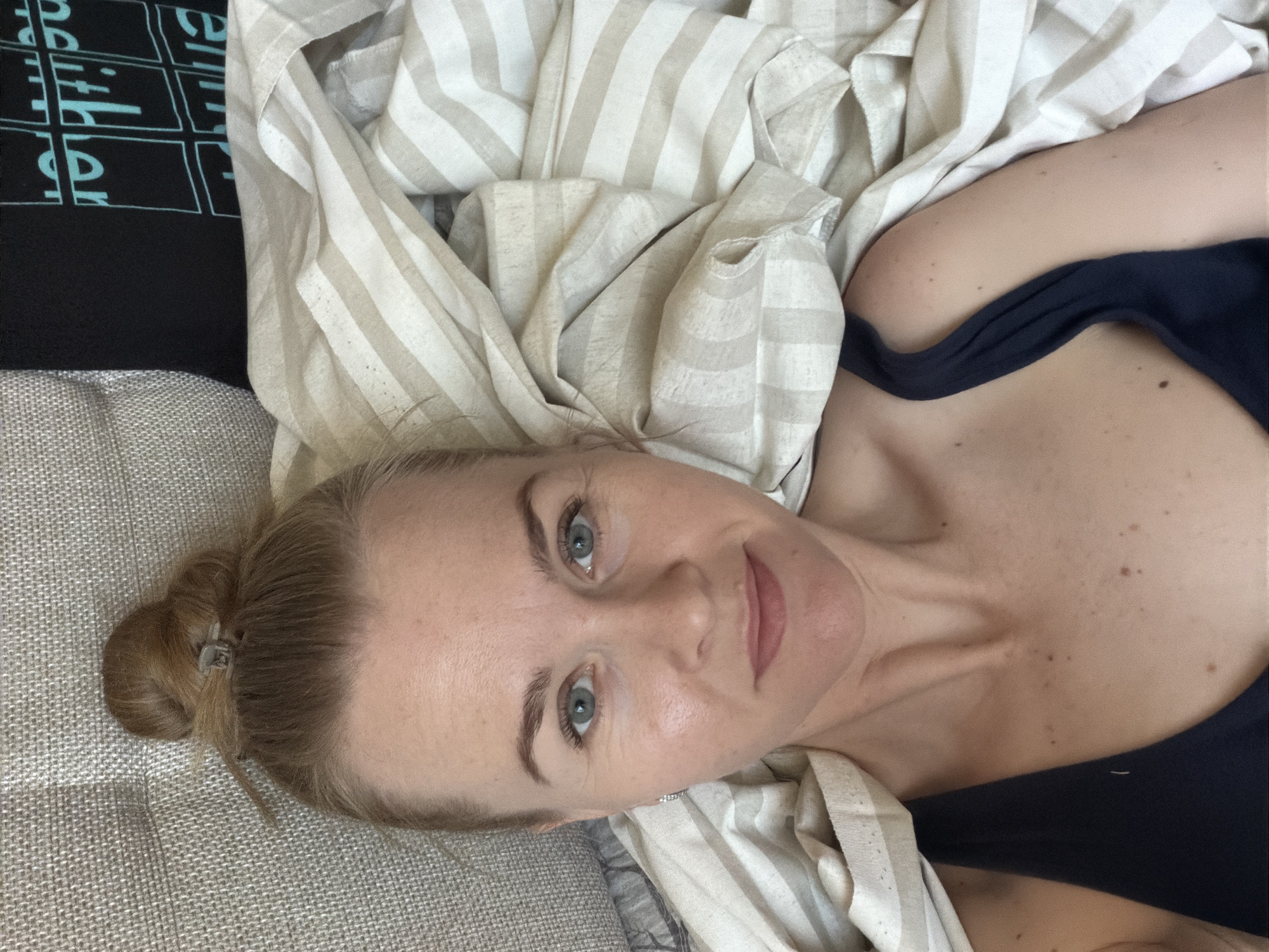 A young woman with her hair in a bun is lying in bed. She is wearing a dark blue tank top. The bed is covered with a striped comforter. The background shows a grey couch and a black t-shirt with blue writing on it. The image is a close-up selfie taken from a high angle. No specific location in Khabarovsk, Russia can be determined from the image.