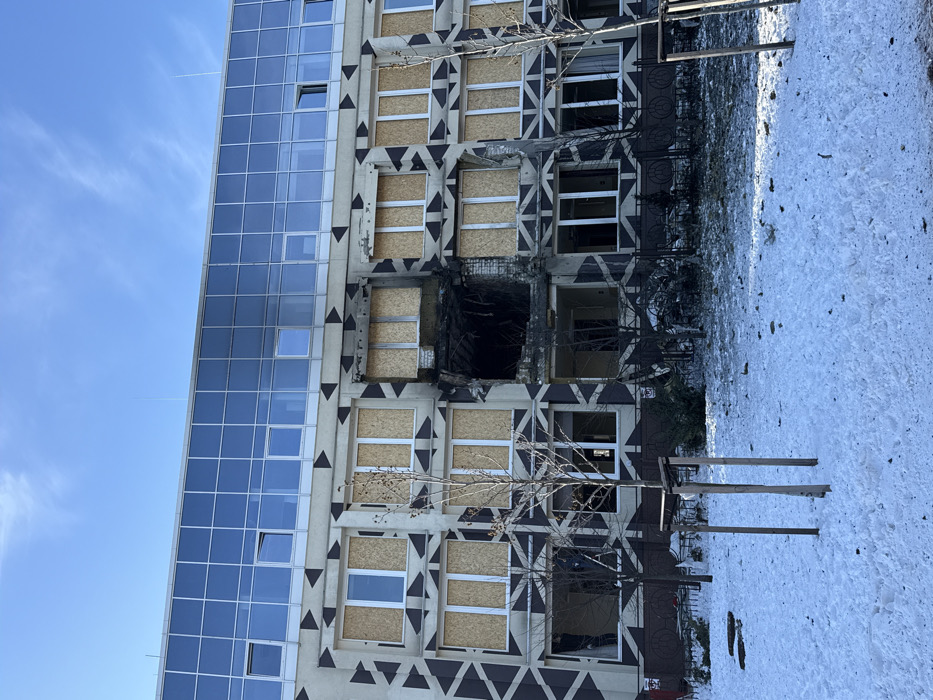 The image shows the Sport Life building at Valeriia Lobanovskoho Avenue, 4A, Kyiv, Ukraine, exhibiting significant structural damage under a clear blue sky. The lower four visible floors of the building feature a light-colored facade with a distinctive dark, geometric arrow pattern. Above these, at least two floors are entirely clad in reflective blue glass paneling. A large, irregular impact hole, blackened by charring and soot, is prominent in the central-right section of the patterned facade, spanning approximately two stories and multiple window bays. Surrounding windows on the damaged floors are either boarded with light-colored plywood panels or are entirely missing glass, revealing dark interiors. Debris is scattered on the thin, patchy snow covering the foreground ground, which is separated from the building by a low, dark metal fence. Several bare deciduous trees stand in the snowy area. No individuals are visible in the scene.
