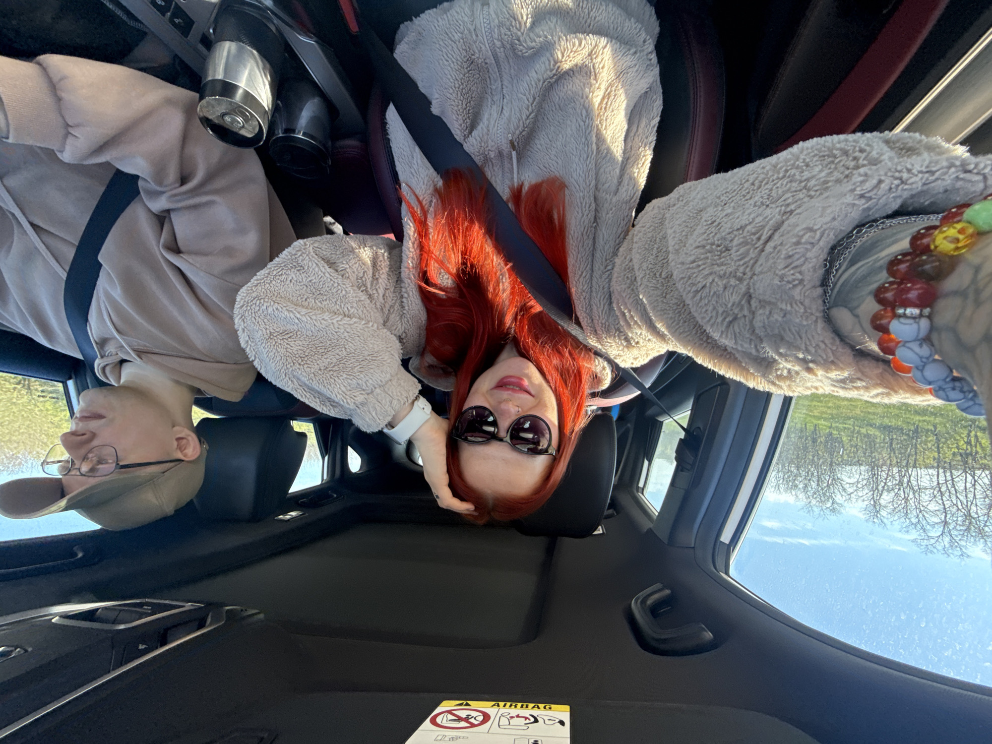 Two individuals are inside a car. A woman with red hair, wearing sunglasses and a fleece jacket, is in the passenger seat. A man with glasses and a baseball cap is in the driver's seat. Both are wearing seatbelts. The car is traveling through a rural area, with trees and sky visible through the windows. An "AIRBAG" label is present on the dashboard. The location may be Pokrovskoye, Russia.