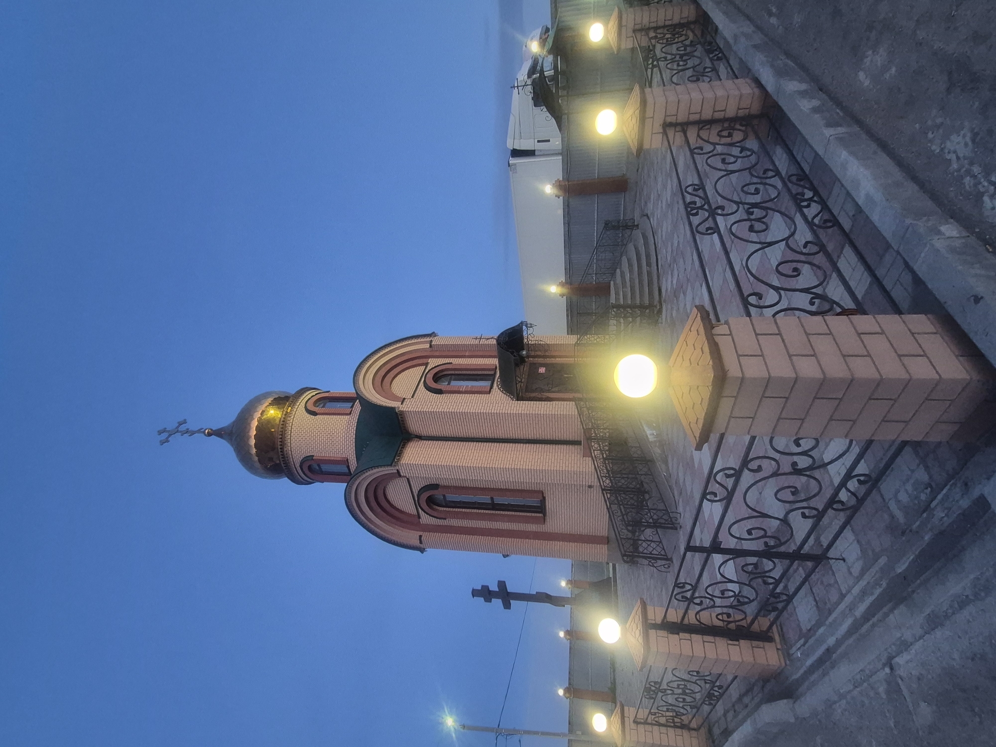 The image depicts the Church of St. Sergius of Radonezh, located in Sergeevka, Russia. The church features a golden dome topped with a cross. The building is constructed of brick with rounded architectural details. In the foreground, there is a decorative wrought-iron fence with brick pillars. Streetlights illuminate the scene. A white truck is parked behind the church. The sky is a deep blue color.