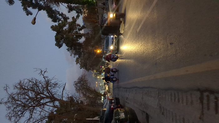 The image depicts an urban street scene at dusk or early night in Quetta City Tehsil, Pakistan. The asphalt road is wet and reflective from artificial light sources, featuring a yellow center line. On the left side, a rough, dark shoulder shows tire tracks, bordered by several parked dark-colored sedans, one with a partial license plate reading "PJ" in the foreground.
In the active traffic lanes, two individuals on separate motorcycles are visible from behind, traveling away from the camera in the left lane. Both wear dark clothing. The second motorcycle from the camera exhibits a red light or reflection at its rear. Traveling towards the camera in the right lane is a light-colored sedan with illuminated headlights, followed by a dark-colored auto-rickshaw with a single illuminated headlight. Additional vehicles with active headlights and taillights are visible further down the street.
The environment is characterized by numerous trees lining both sides of the road; many are bare-branched, while others are evergreen. Streetlights are illuminated, with one prominent fixture visible on the upper right. Distant mountains or hills are faintly silhouetted against a blue-grey, cloudy twilight sky. Buildings are partially visible behind the trees in the background.