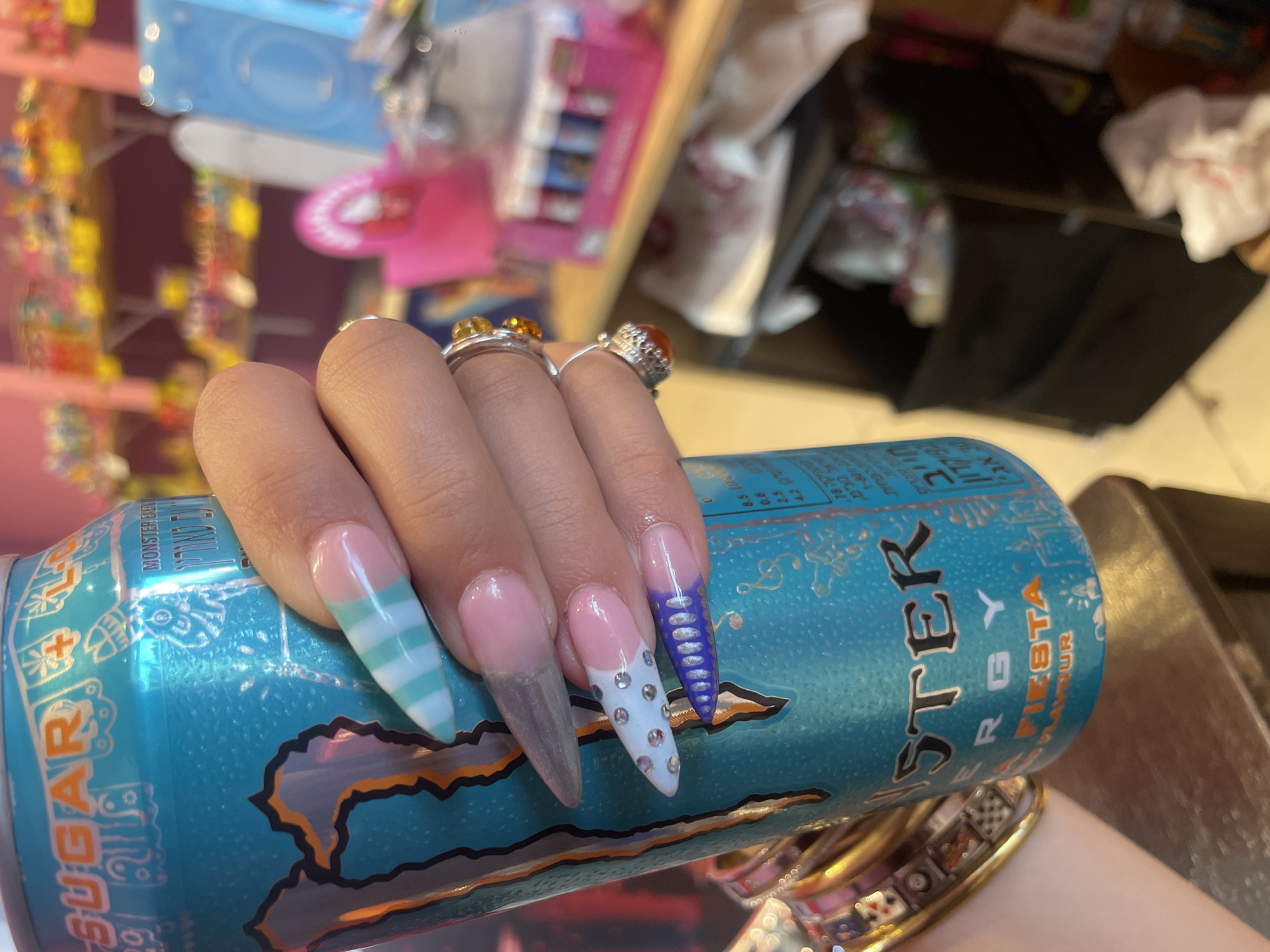 A close-up shot captures a person's hand adorned with elaborate, brightly colored acrylic nails, holding a can of Monster Energy drink. The nails feature a variety of designs, including pastel stripes, French tips with embellishments like rhinestones and metallic detailing, and a grey, pointed nail. The Monster Energy can is a vibrant turquoise color with its distinctive logo and text. The background is out of focus but suggests an indoor environment with shelves stocked with colorful items, possibly a store or a candy shop. The overall impression is one of a vibrant and playful aesthetic. No specific landmarks or identifiable features of Raanana, Israel, are visible in the image.