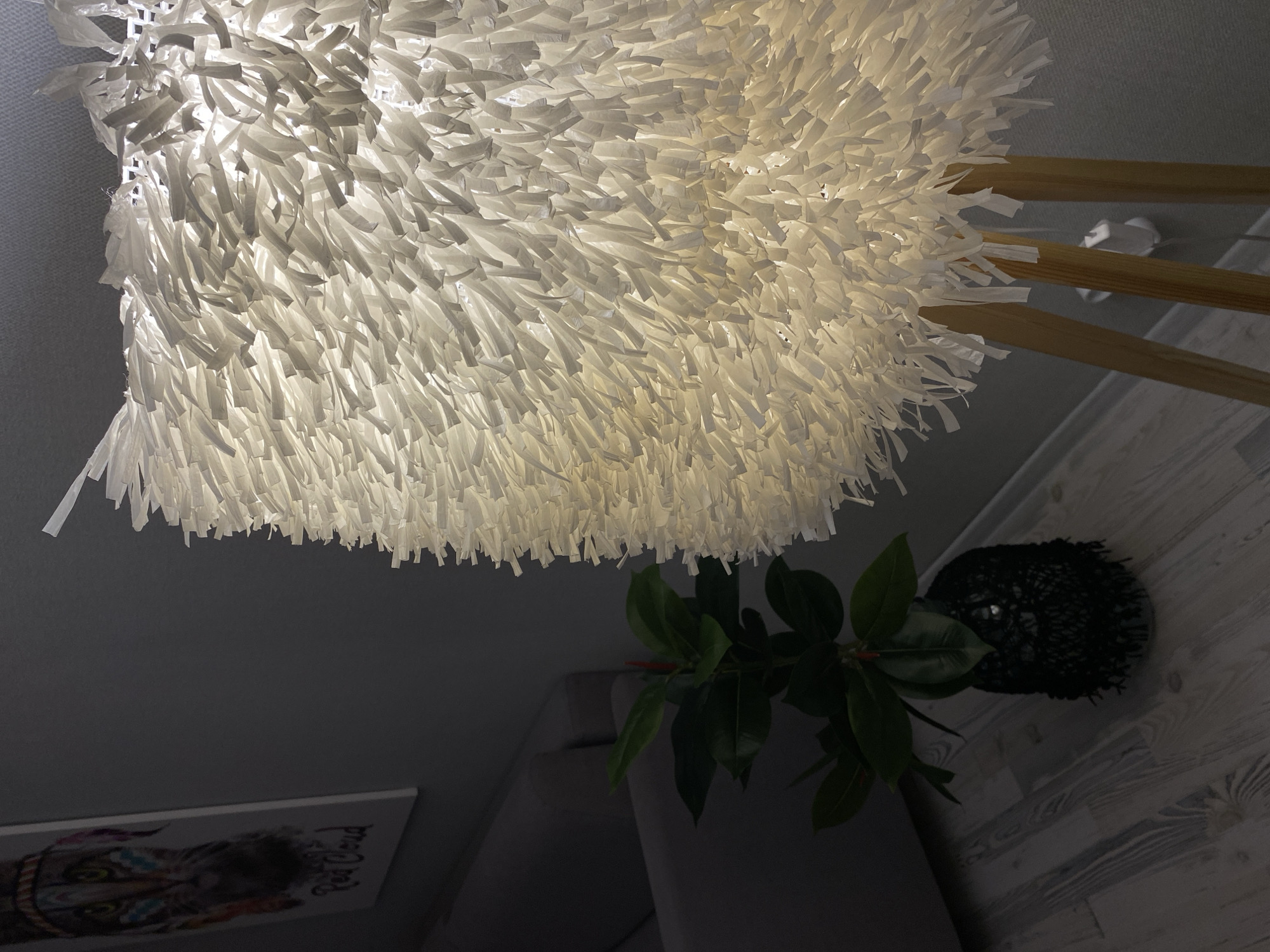 This is a close-up, indoor shot of a lamp with a textured, white lampshade made of frayed strips of material. The lamp has a wooden tripod base. To the left of the lamp is a plant in a black pot, and further left is a grey sofa and a framed picture of a tiger's face with Native American-inspired face paint. The floor is made of light-colored wood planks. The image does not contain any information that allows for the determination of its location in Votkinsk, Russia.
