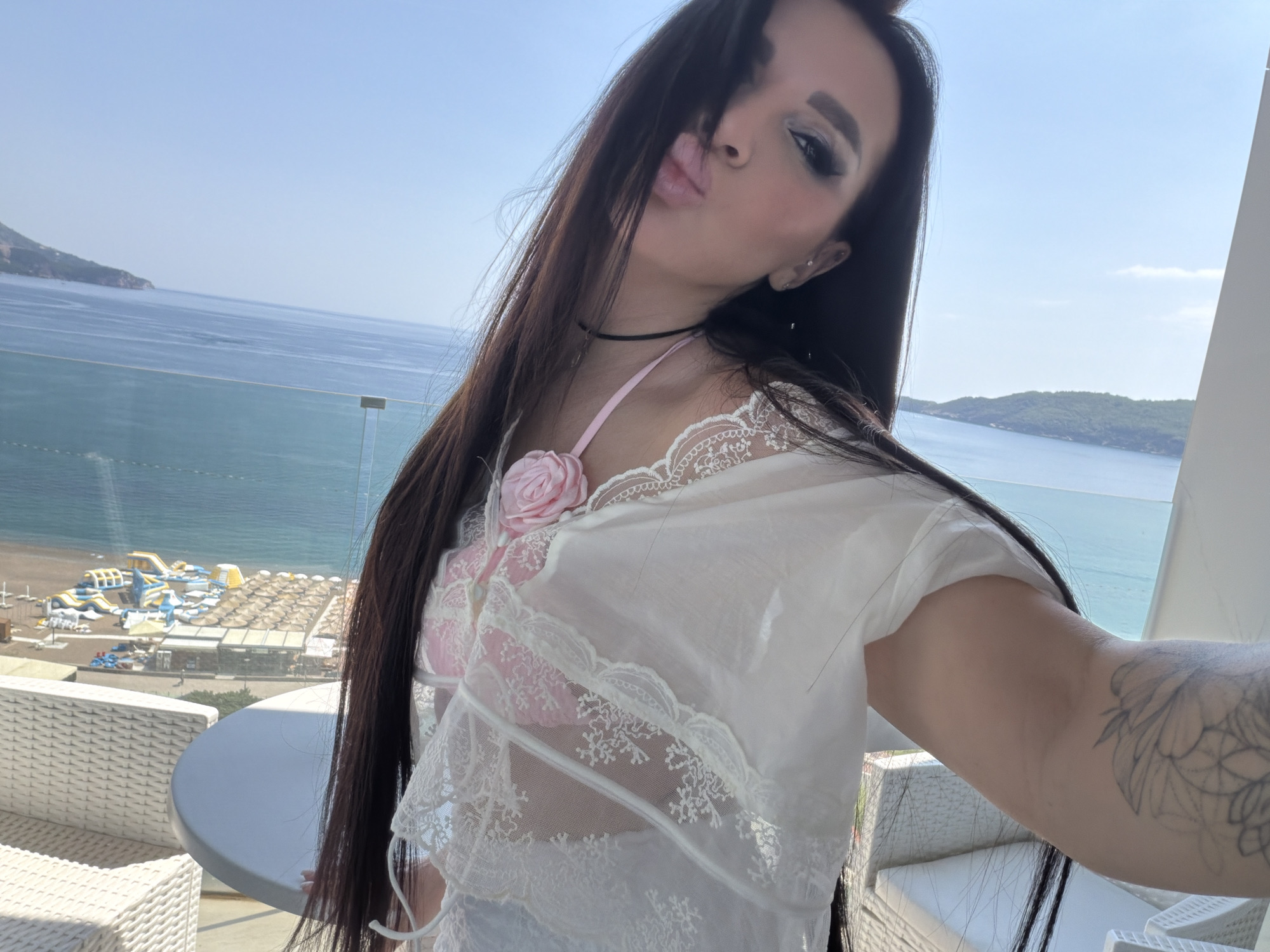 A fair-skinned woman with long, dark hair is taking a selfie outdoors. She is wearing a white, lace-trimmed top over a pink garment. She has a black choker necklace. Her left arm is visible, revealing a tattoo on her forearm. The background shows a coastal scene with light blue water and a beach. On the beach are rows of lounge chairs and sun umbrellas, along with inflatable water park structures. The location is likely a balcony or elevated position overlooking the beach. This is Boreti, Montenegro.