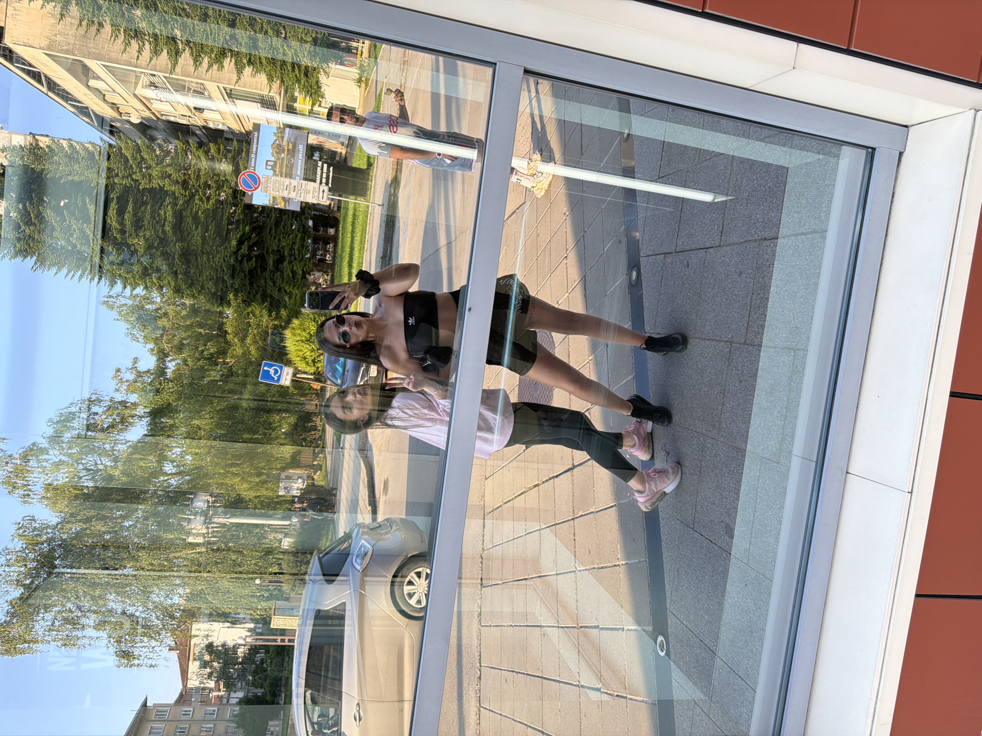 This image is a reflection captured in a large glass pane, likely a window or bus stop shelter, depicting an urban street scene in Gabrovo, Bulgaria, during daytime.
In the foreground of the reflection are two young females posing. The female on the left, with long dark hair, is wearing a light pink long-sleeved top, dark leggings, and pink athletic shoes. She is making a "peace" sign gesture with her right hand and pouting her lips, looking towards the camera. The female on the right, also with long dark hair tied up, is wearing sunglasses, a black strapless crop top (with an Adidas logo visible), black shorts or a skirt, and black athletic shoes. She is holding a black smartphone in her right hand, appearing to take a selfie. A small black purse hangs from her left shoulder.
In the mid-ground reflection, on the sidewalk behind the two females, a man is visible. He has dark hair, is wearing a white t-shirt with a graphic (possibly "220") and light-colored shorts, and is holding an ice cream cone in his right hand.
The background reflection includes a street with parked vehicles, green spaces, and buildings. A light-colored (possibly silver) 5-door hatchback is parked on the left side of the street. Further to the right, a dark-colored car is partially visible. Numerous green trees, both deciduous and coniferous, are reflected, suggesting a park or tree-lined area. Street furniture includes a blue sign with a wheelchair symbol, a red circular "no entry" or "no stopping" sign, and a large advertising or information board displaying Cyrillic text, including "Mapeh.AG" and phone numbers (e.g., "0 88 49 6350"). Various multi-story buildings are reflected, exhibiting different architectural styles, from older residential structures on the left to more contemporary or brutalist designs on the right. A traditional street lamp post is also visible. The ground is paved with concrete tiles, showing patterns of light and shadow from the sun.
The photograph itself captures the glass pane, showing a light gray frame around the edges and a horizontal crossbar in the middle. The bottom right corner of the actual frame reveals reddish-brown paneling with white trim, indicating the structure from which the reflection is being observed.
