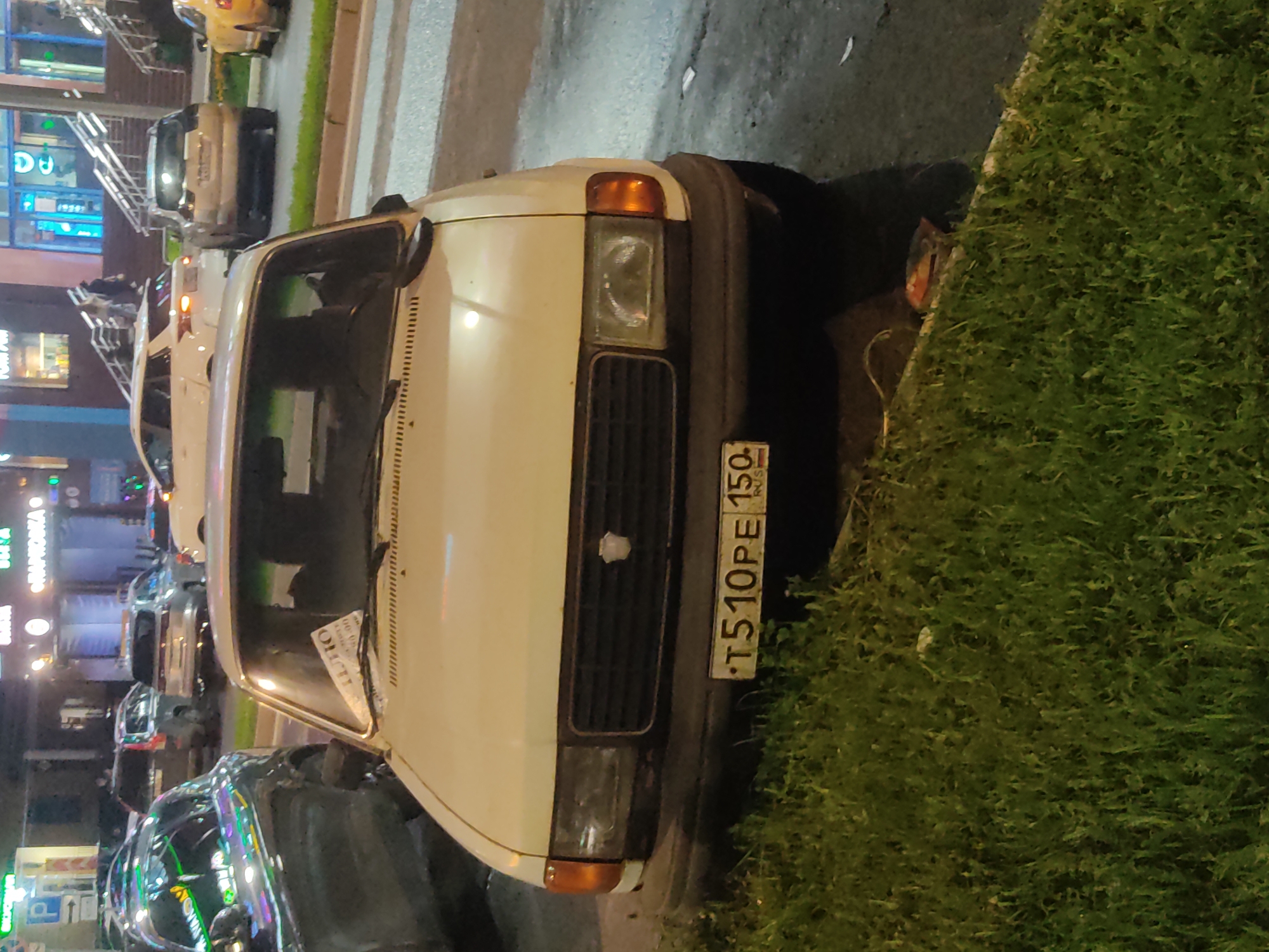 The image shows the front of a white car parked near a grassy area on a city street. The car has a visible license plate reading "T510PE 150". Other cars are visible in the background, parked along the street. Buildings and streetlights suggest an urban setting at night. There is pedestrian crossing behind the car.
