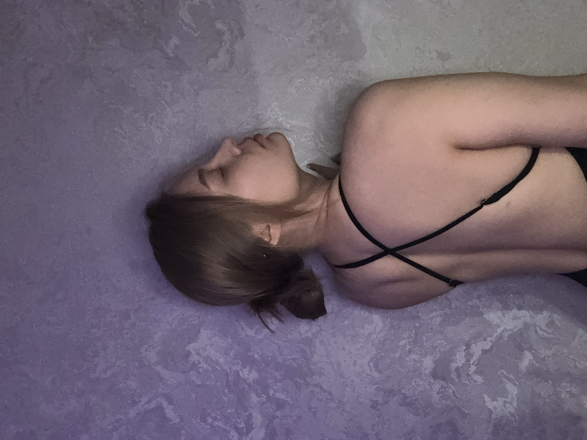 A person is depicted lying on their side on a textured surface. The individual, appearing to be female, has light brown hair gathered into a low bun or ponytail. Her head is turned slightly to the right, presenting a profile view, with her eyes closed or cast downwards, suggesting a state of rest or contemplation. She is wearing a black garment, possibly a top or swimsuit, featuring thin straps that crisscross across her bare upper back. A small, delicate earring is visible in her left ear.
The background is a wall or floor with a distinct marbled or mottled pattern, predominantly in shades of lavender, purple, and lighter grey or white. The lighting is soft and subdued, creating a serene and calm atmosphere. No specific time of day or weather conditions can be discerned from the indoor setting. There are no identifiable location cues within the image itself, nor is any text visible. The overall mood conveyed is one of tranquility and repose.