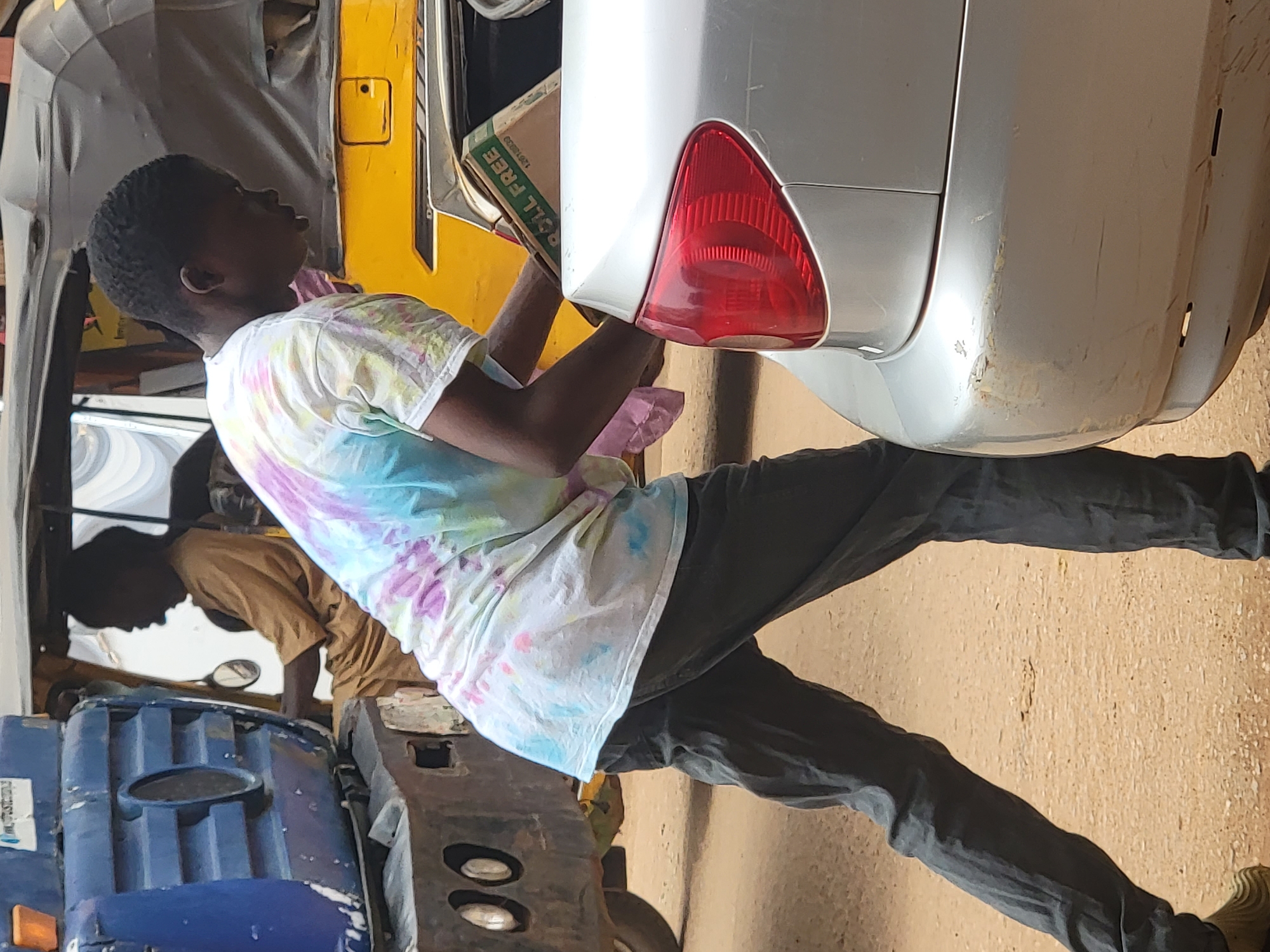 In Maiburiji, Nigeria, a young man in a tie-dye shirt and gray pants stands beside a silver car, holding a cardboard box. He appears to be interacting with the vehicle. The environment includes a blue auto rickshaw, a yellow tuk-tuk, and other individuals in the background, all situated on a paved surface. The overall scene suggests an urban or roadside setting.
