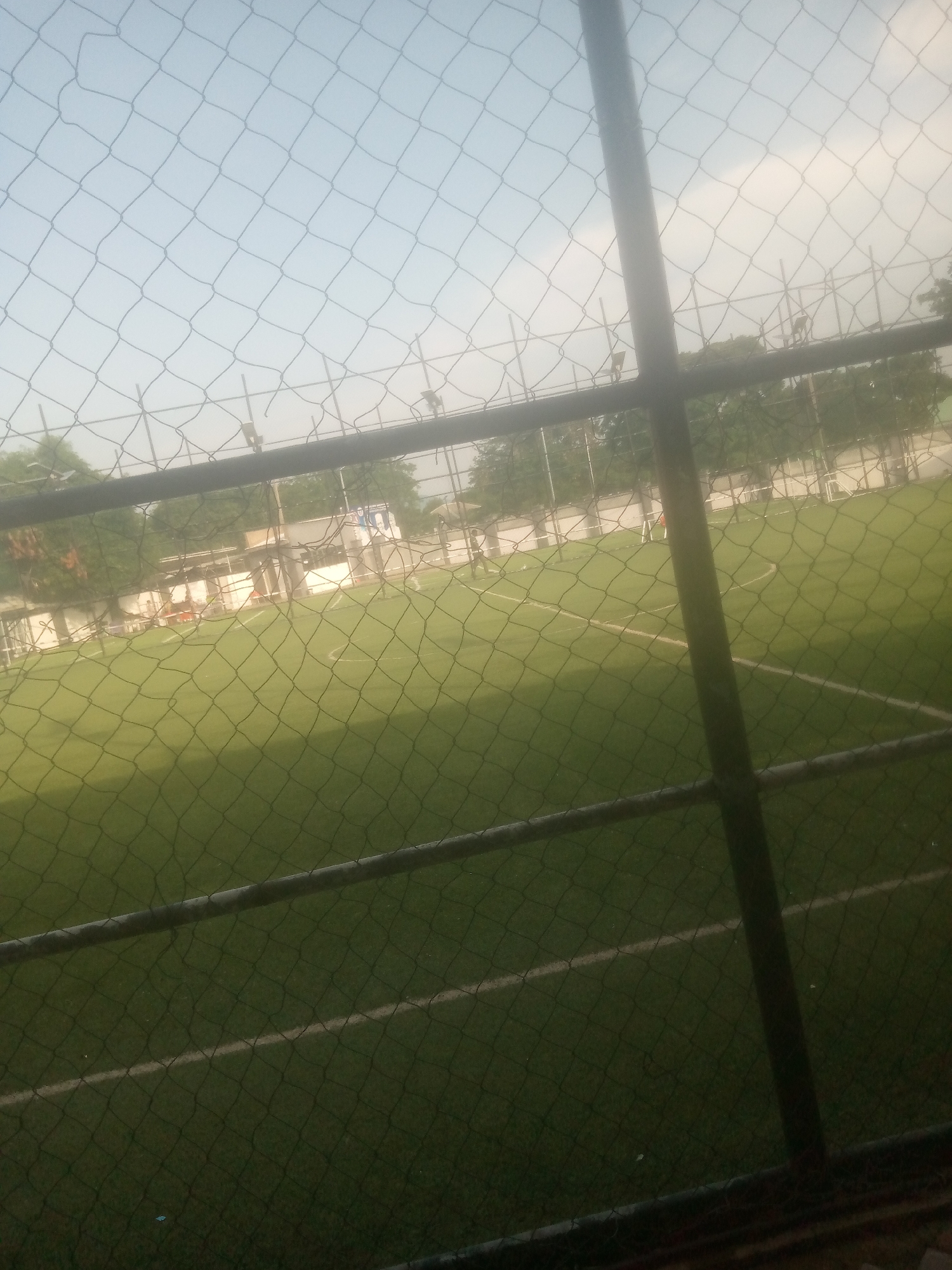 The image depicts an outdoor football pitch, likely artificial turf, viewed through a chain-link fence. The sky is a pale blue with some hazy clouds, suggesting it is daytime, possibly late afternoon due to the soft light. Trees and some low buildings are visible in the background, behind the pitch and a further fence. The football pitch has white lines marking its boundaries and possibly a center circle. There are a few people visible on the pitch, though they are small and indistinct due to the distance and the fence obstruction. They appear to be participating in a game or training session. The overall scene suggests a recreational or sporting activity taking place in a somewhat overcast but clear environment. There is no visible text in the image. The location context of Jalingo, Nigeria, would place this scene in West Africa.