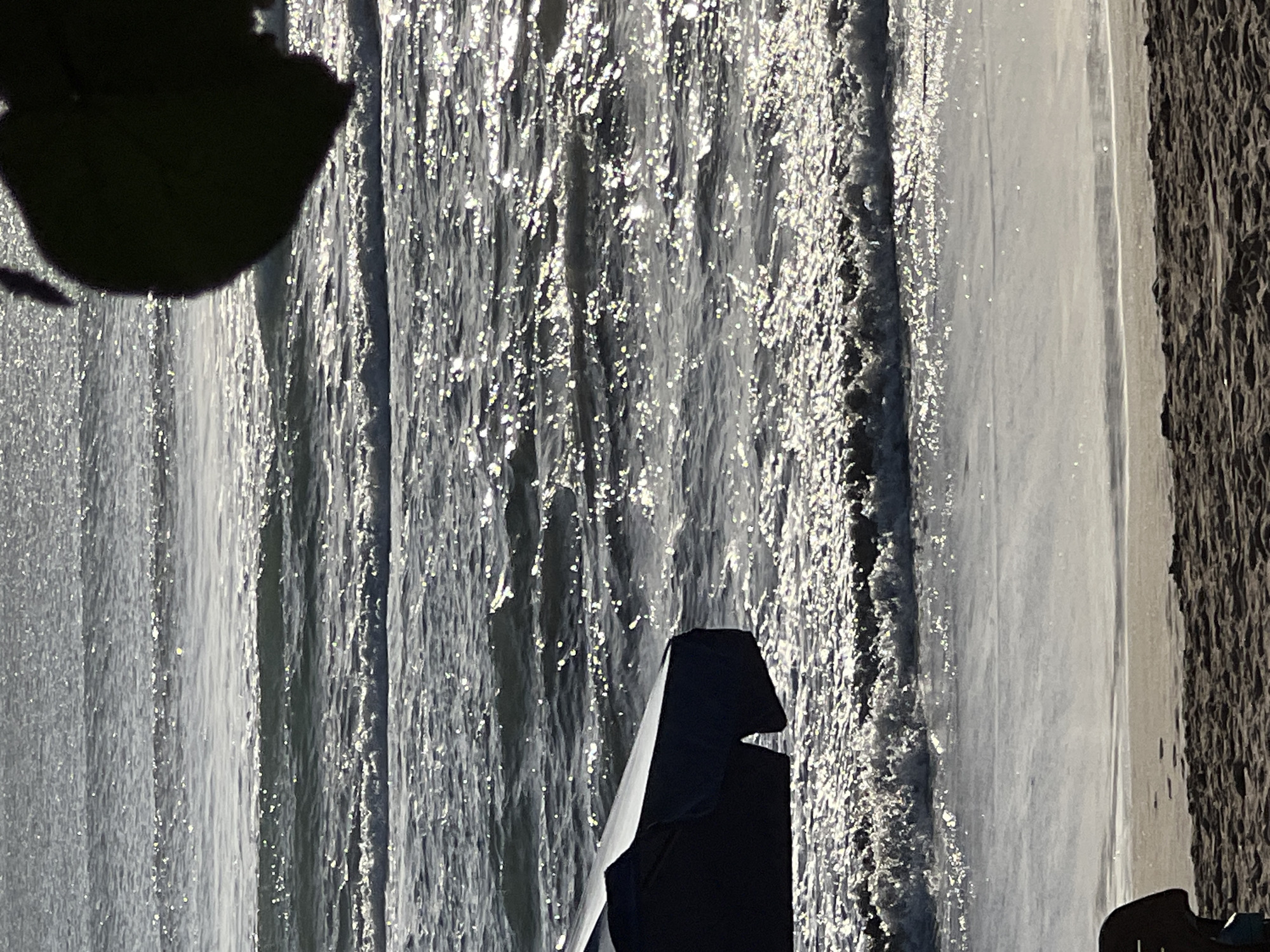 The image depicts a sun-drenched beach scene with the ocean and sand. In the foreground, a dark blue beach umbrella is partially visible on the left, angled towards the sea. The sand is coarse and textured, with some ripples. The ocean is choppy, with waves rolling towards the shore, reflecting the sunlight and creating a shimmering effect. The water is a dark grey-blue, with white foam on the breaking waves. In the upper right corner, a dark silhouette of a leaf or foliage obscures a portion of the scene. The overall impression is one of a bright, breezy day at the coast. The image is consistent with a scene found in Kuta, Indonesia, a popular tourist destination known for its beaches and surf.