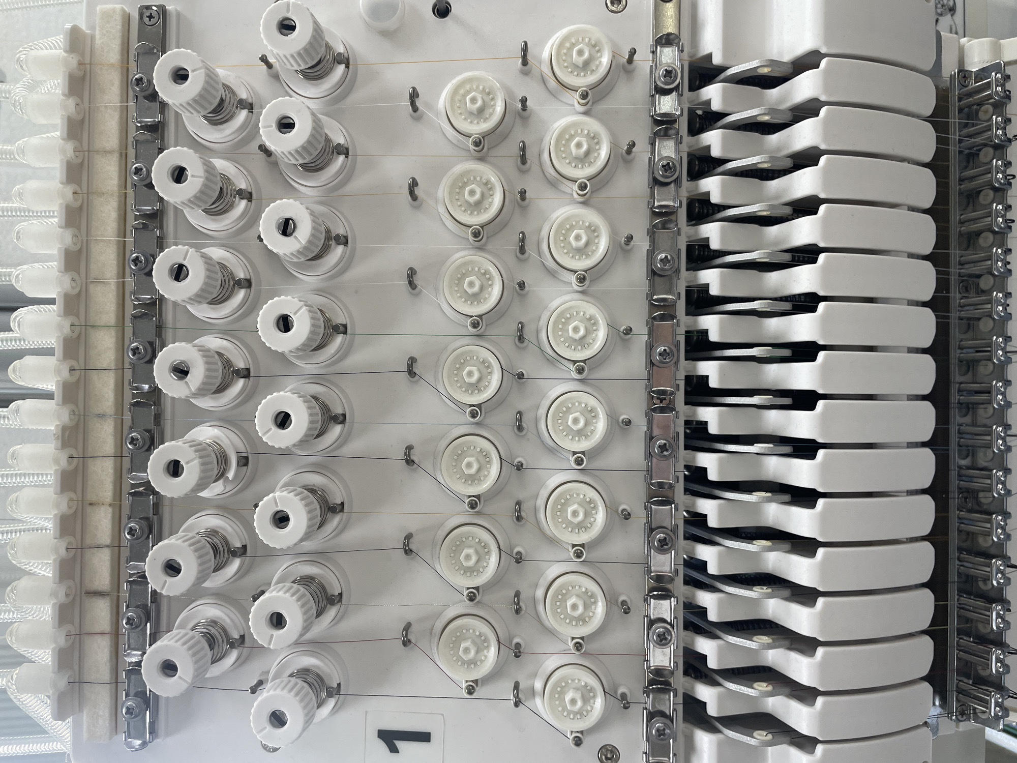 A close-up, eye-level, interior shot shows a section of an embroidery machine's thread spool and needle area, set against a neutral white background. The focus is on the organized arrangement of spools and the intricate threading system.
The top portion of the image displays a row of white, cylindrical thread spools, each with a textured, ribbed cap. These spools are mounted on a white plastic rack, with individual threads running down from them. Below the spools, a metal bar with several screw heads serves as a guide for the threads.
Further down, two rows of white, hexagonal spool holders are visible, each with a thread passing through its center. The threads, in various colors including red, yellow, and green, are meticulously guided through small metal eyelets on the machine.
The bottom section of the image reveals a row of white, U-shaped thread guides, positioned above a series of silver metal needle holders. Each needle holder appears to house a needle, with threads ready to be fed through. The overall impression is one of precision and mechanical complexity. The image is devoid of any human presence, signs of wear, or damage, suggesting a well-maintained piece of equipment. There is no indication of its location in a city or geographical context.