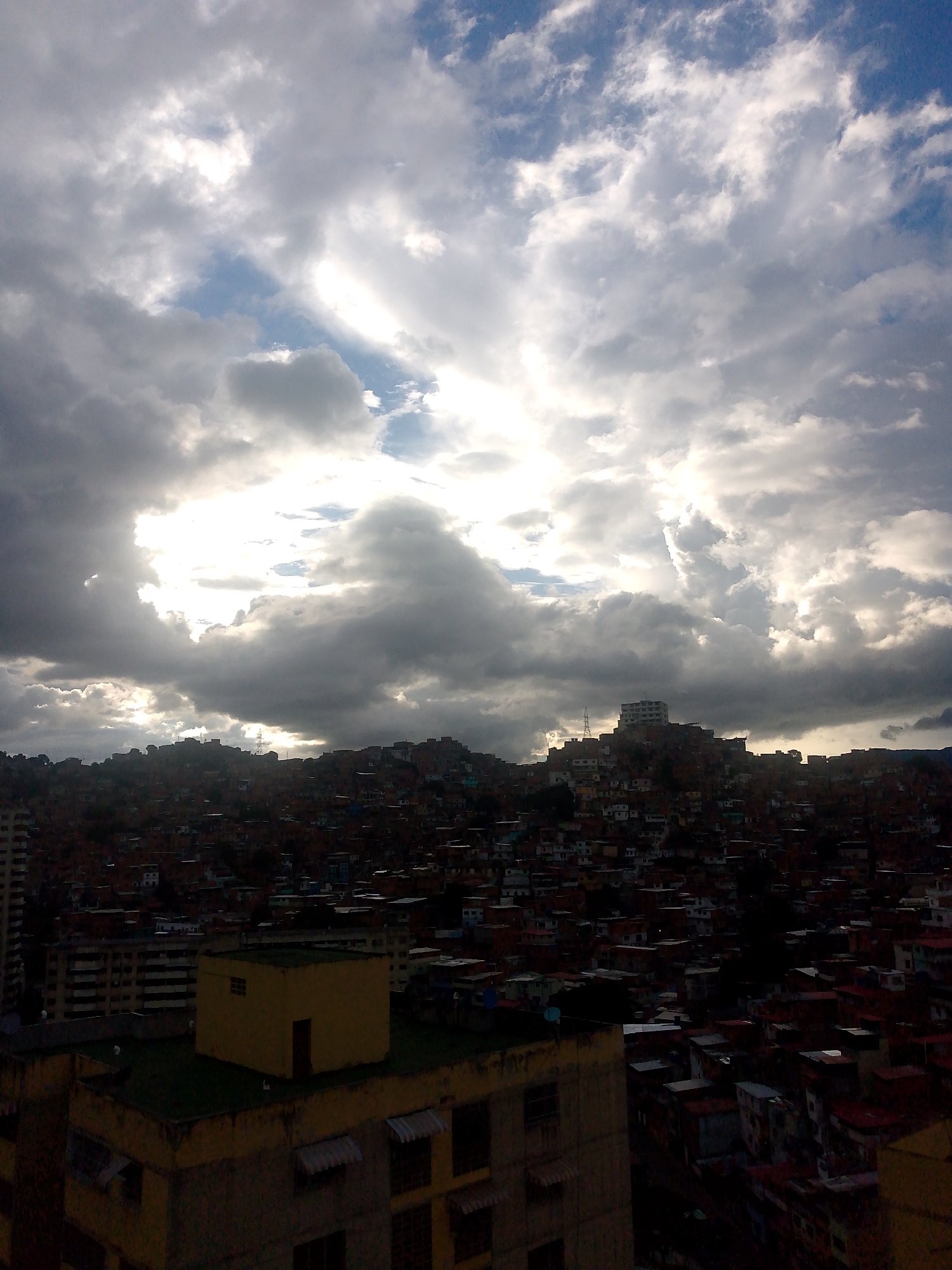 The image depicts a dense urban hillside cityscape in Caracas, Venezuela, under a dramatic sky filled with heavy, gray clouds partially illuminated by the sun. The densely packed buildings, characterized by their red-tiled roofs and various colors, cascade down the steep terrain, indicative of a barrio or informal settlement. In the foreground, several concrete buildings are visible, some appearing to be multi-story residential structures or apartment complexes. One prominent building in the foreground is a lighter colored structure topped with a small, square, yellow building with a single door and a small window. The background is dominated by the vast, turbulent sky, with bright patches where the sun is breaking through the clouds, creating shafts of light that illuminate parts of the city. The overall impression is one of a sprawling urban environment set against a powerful and dynamic natural backdrop. There are no people visible in the image.