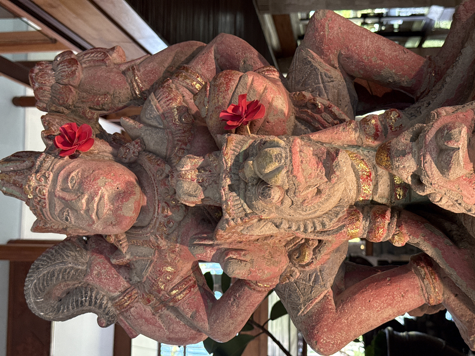 The image shows a close-up, vertical shot of a weathered, carved stone statue of a multi-limbed deity, possibly from a Balinese temple, located in Kuta Selatan, Indonesia. The statue is predominantly red with gray accents, and features gold detailing on its headdress, jewelry, and armor. Two small, red artificial flowers are affixed to the statue, one near its headdress and another on its chest. The deity is depicted in a seated, dynamic pose, with multiple arms positioned in various gestures, holding what appears to be a sword or scepter. It sits atop a smaller, monstrous creature that is also carved from stone and shares similar coloration and decorative elements. The background is blurred, but suggests an indoor setting with wooden architectural elements and hints of greenery visible through glass. The lighting appears to be a mix of natural and artificial sources, casting subtle shadows and highlighting the texture of the stone.