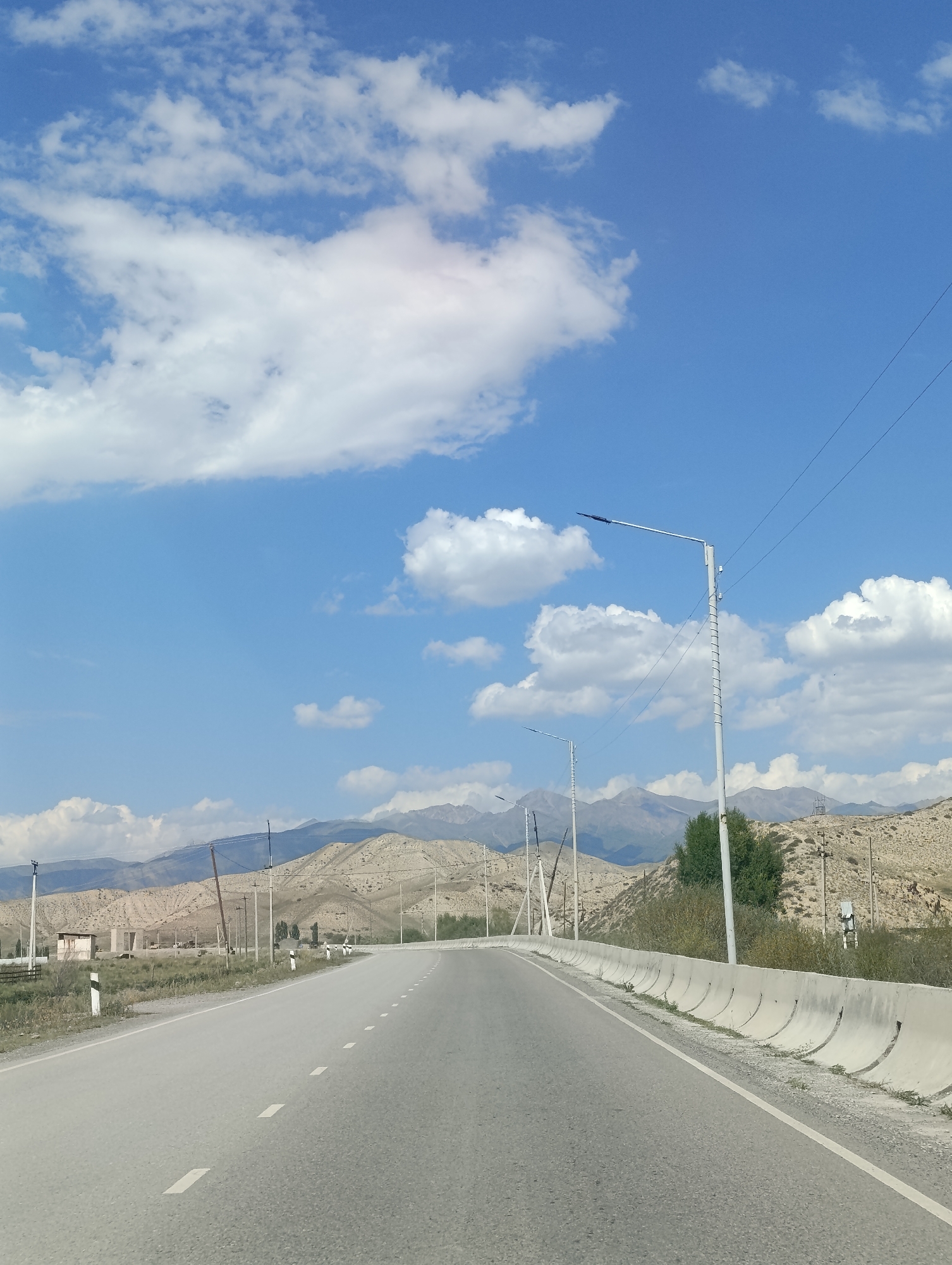 The image depicts a road leading into a mountainous landscape under a bright blue sky with scattered white clouds. The road is paved with asphalt and has white dashed lines marking the lanes. On the right side of the road, there is a concrete barrier and a lamppost. In the distance, hills and mountains are visible, with sparse vegetation and some structures. The scene appears to be an exterior shot on a sunny day, possibly in a rural area or on the outskirts of a town. The location is specified as Naryn, Kyrgyzstan.