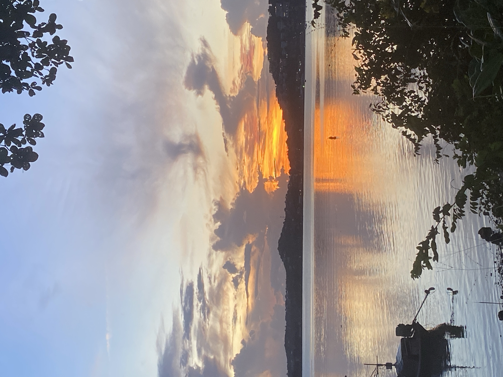 The image depicts a serene sunset over a body of water in Baan Bang Rak, Thailand. The sky is filled with a gradient of colors, transitioning from a pale blue at the top to warm oranges and pinks near the horizon, accented by dramatic clouds. The calm water reflects the vibrant sky, creating a mirror-like effect.
In the foreground, a dark silhouette of a small boat with an outboard motor is partially submerged near the shore, its reflection visible in the water. Lush green foliage, also silhouetted, frames the right side of the image, adding depth and a natural element. Across the water, a dark, hilly landscape is visible, dotted with lights that suggest a settlement or town. A lone figure is standing in the shallow water near the center of the frame, their silhouette small against the expansive sunset. The overall scene conveys a sense of tranquility and natural beauty.
