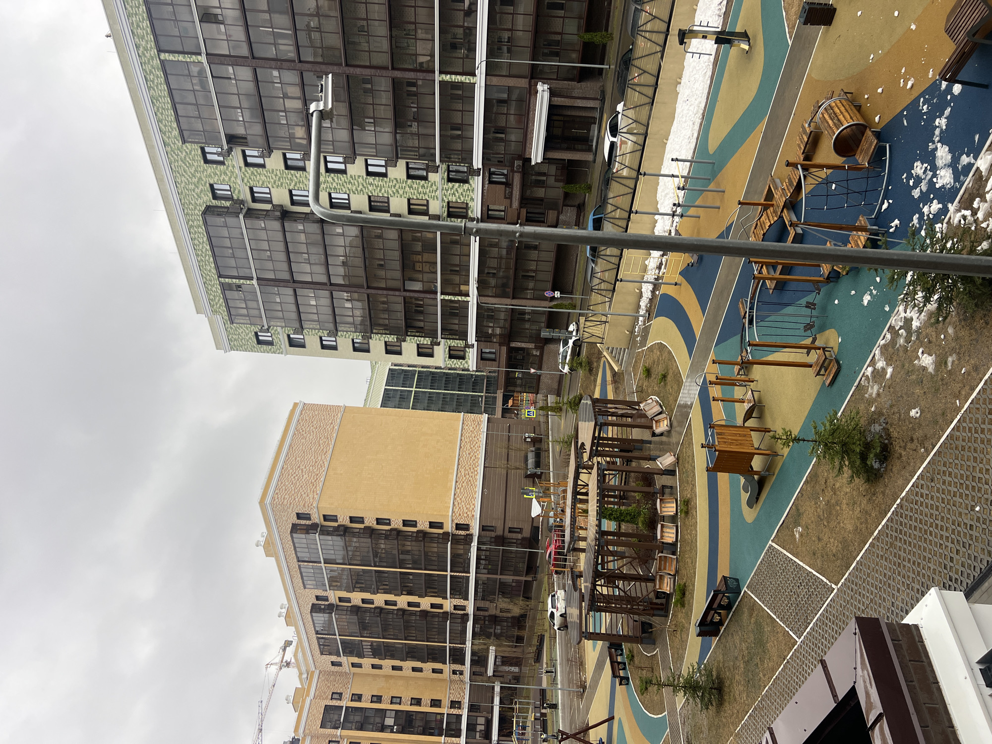 The image depicts a residential area in Kazan, Russia, featuring modern apartment buildings and a playground. Two multi-story buildings with distinct architectural styles are visible. The building on the left has a yellowish facade and multiple rows of windows. The building on the right exhibits a green and gray patterned facade with similar window arrangements. A playground is located between the buildings, with colorful ground surfaces and wooden play structures. The playground shows signs of use, and some areas are covered in snow. A street lamp is visible in the foreground. The sky is overcast, suggesting a cloudy day.