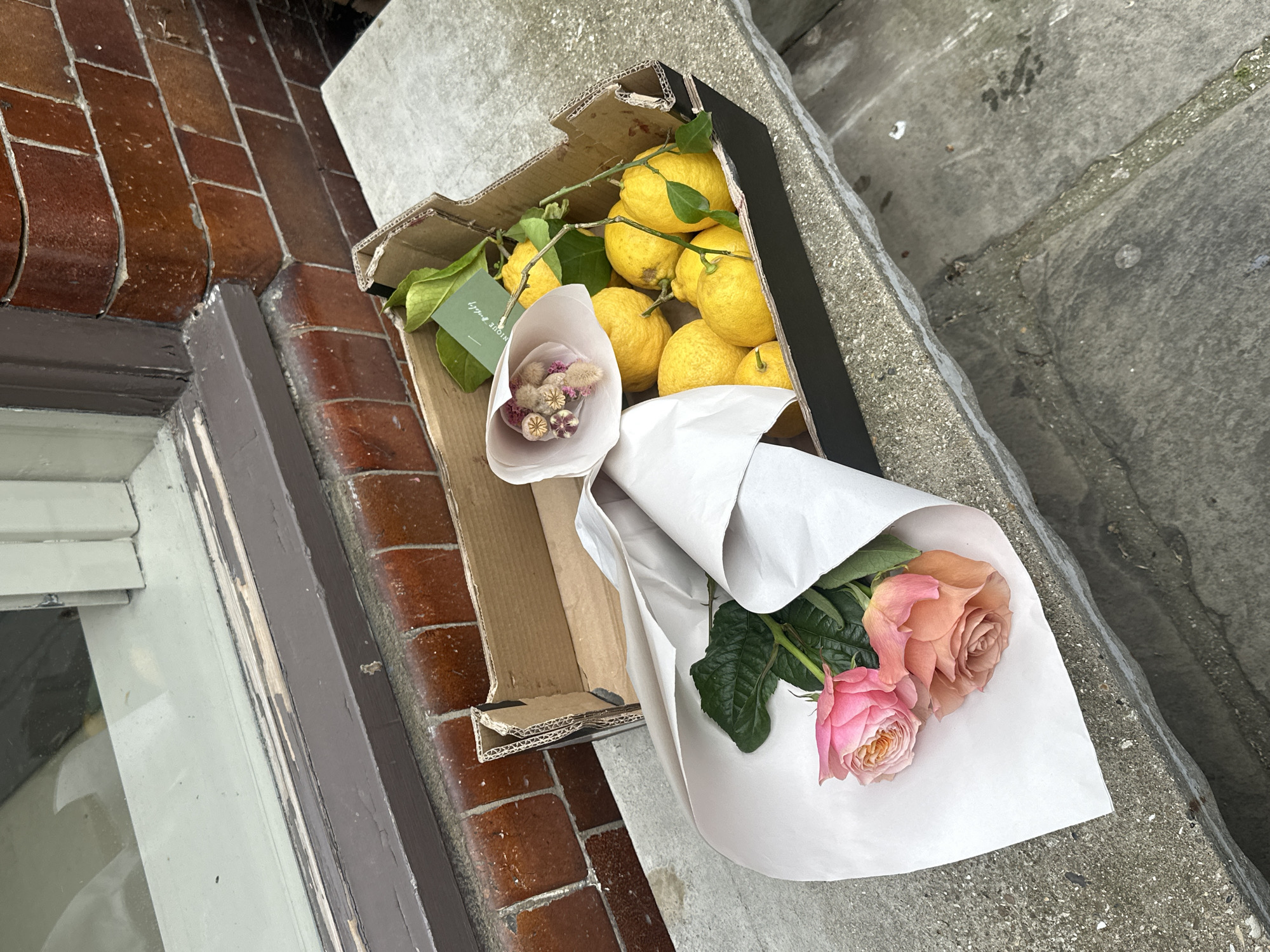 A cardboard box filled with lemons, a bouquet of dried flowers, and a bouquet of roses sits on a concrete step outside a building in London, United Kingdom. The box is positioned at an angle, partially on the step and partially on brickwork. The lemons have green leaves still attached. One bouquet is small and contains dried flowers wrapped in white paper. The other bouquet contains roses wrapped in white paper, with visible pink and peach blooms. The background includes part of a window and brick wall on one side, and the concrete step continuing upwards on the other.