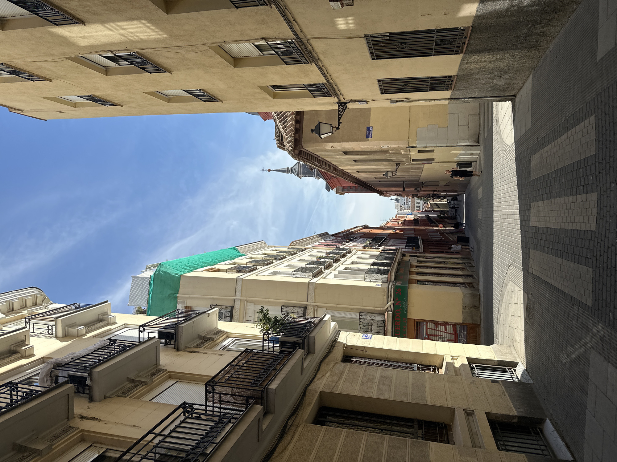 The image shows a narrow street in Madrid, Spain, looking upward. The street is lined with buildings of various heights and styles, ranging from traditional to more modern architecture. The buildings are primarily light beige or yellow. Balconies with black iron railings are visible on some of the buildings. A green tarp is covering part of one building. At the end of the street, a steeple is visible in the distance. The sky is blue with light clouds. The street is paved with light and dark gray paving stones, and a sidewalk runs along the right side. A few pedestrians are visible further down the street. A street sign that says "Manuel" is on one building.