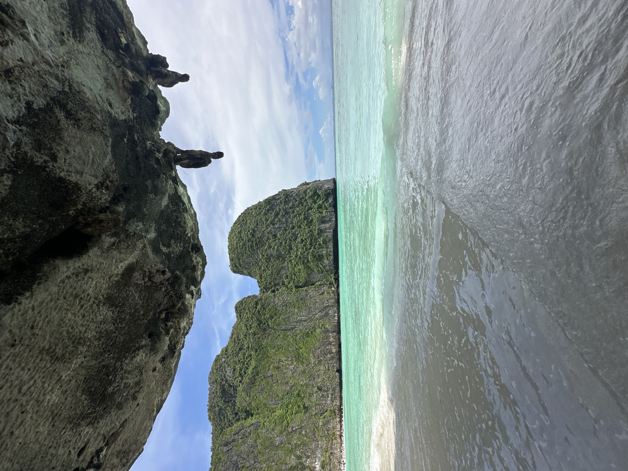The image depicts a tropical beach scene with dramatic limestone cliffs framing the view. In the foreground, a large, rough-textured grey rock juts out from the left, its surface showing signs of erosion and moss growth. Above this rock, silhouetted figures appear to be perched on the cliff edge, adding a sense of scale and human presence.
The middle ground is dominated by a lush, green, jungle-covered karst island rising steeply from the turquoise waters. The island has a rugged, uneven texture, characteristic of these geological formations.
To the right of the island, the ocean stretches out, with clear, shallow waters transitioning into deeper, darker hues. A gentle wave is breaking near the shore, its crest catching the light. The beach itself is visible as a narrow strip of pale sand. In the distance, along the waterline at the base of the karst island, a line of people can be seen, suggesting this is a popular tourist destination.
The sky above is a bright blue, dotted with wispy white clouds, indicating fair weather. The lighting suggests it is daytime, likely late morning or early afternoon. The overall scene evokes a feeling of natural beauty and tranquility, with the presence of people hinting at a vibrant, accessible location. The image is oriented vertically, with the sky at the bottom and the sea at the top, giving a unique perspective. There is no visible text in the image. The location is identified as Ao Nang, Thailand, which is known for its stunning karst landscapes and beaches.