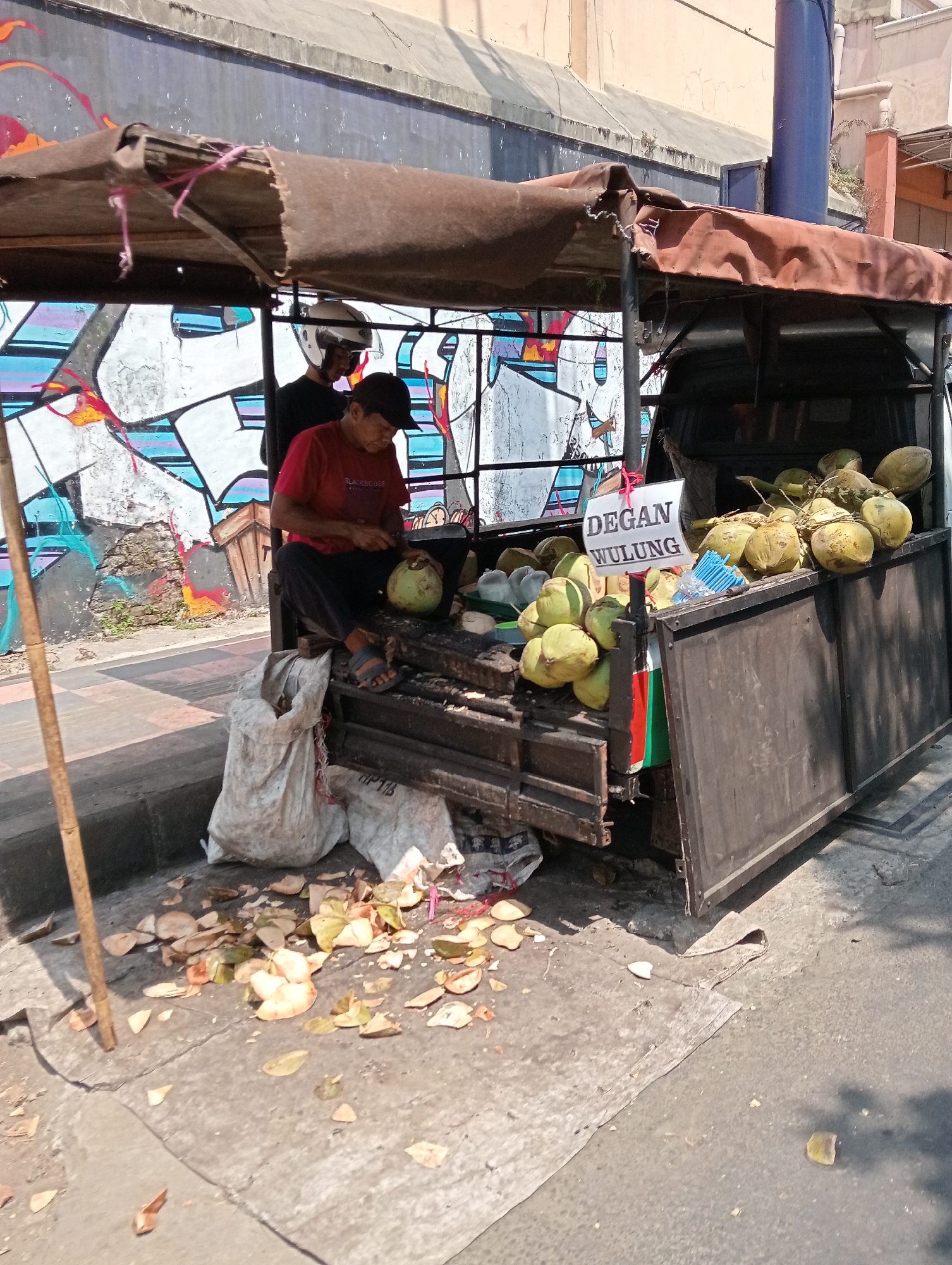 The image shows a street vendor in Grabag, Indonesia, selling coconuts from a cart. A man in a red shirt and black pants is sitting on the cart, peeling a coconut with a knife. Another person wearing a white helmet is standing behind him. The cart is loaded with fresh coconuts, and there is a sign that reads "DEGAN WULUNG". The ground around the cart is littered with coconut husks and leaves. In the background, there is a colorful mural on a wall. The scene is illuminated by bright sunlight, casting shadows on the ground.