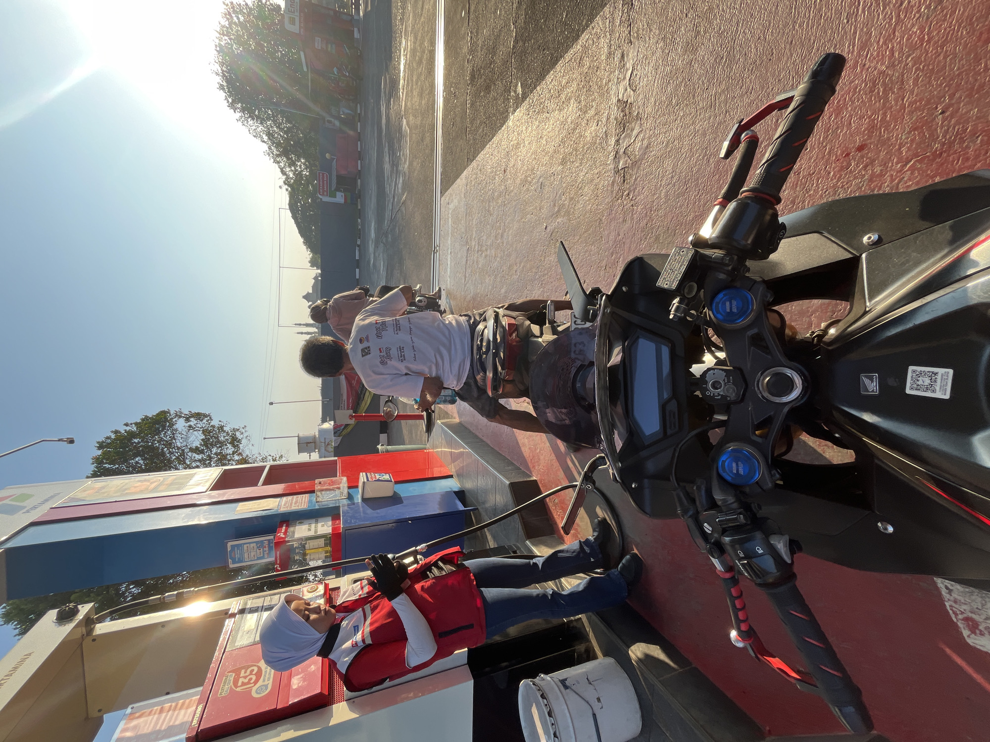 The image depicts a petrol station in Pecangaan, Indonesia, during daylight hours, possibly in the morning due to the low angle of the sun. The foreground is dominated by the handlebars and front of a black motorcycle, suggesting the photo is taken from the rider's perspective. In the mid-ground, a female attendant wearing a red vest and a light blue hijab is standing at a Pertamina gas pump, holding a fuel nozzle. She appears to be in the process of refueling a motorcycle. Two other individuals on motorcycles are also visible, one in front of the attendant and another slightly further back. The environment consists of a paved area typical of a petrol station, with gas pumps, signage, and trees in the background. The sky is clear and bright with the sun creating a lens flare effect.