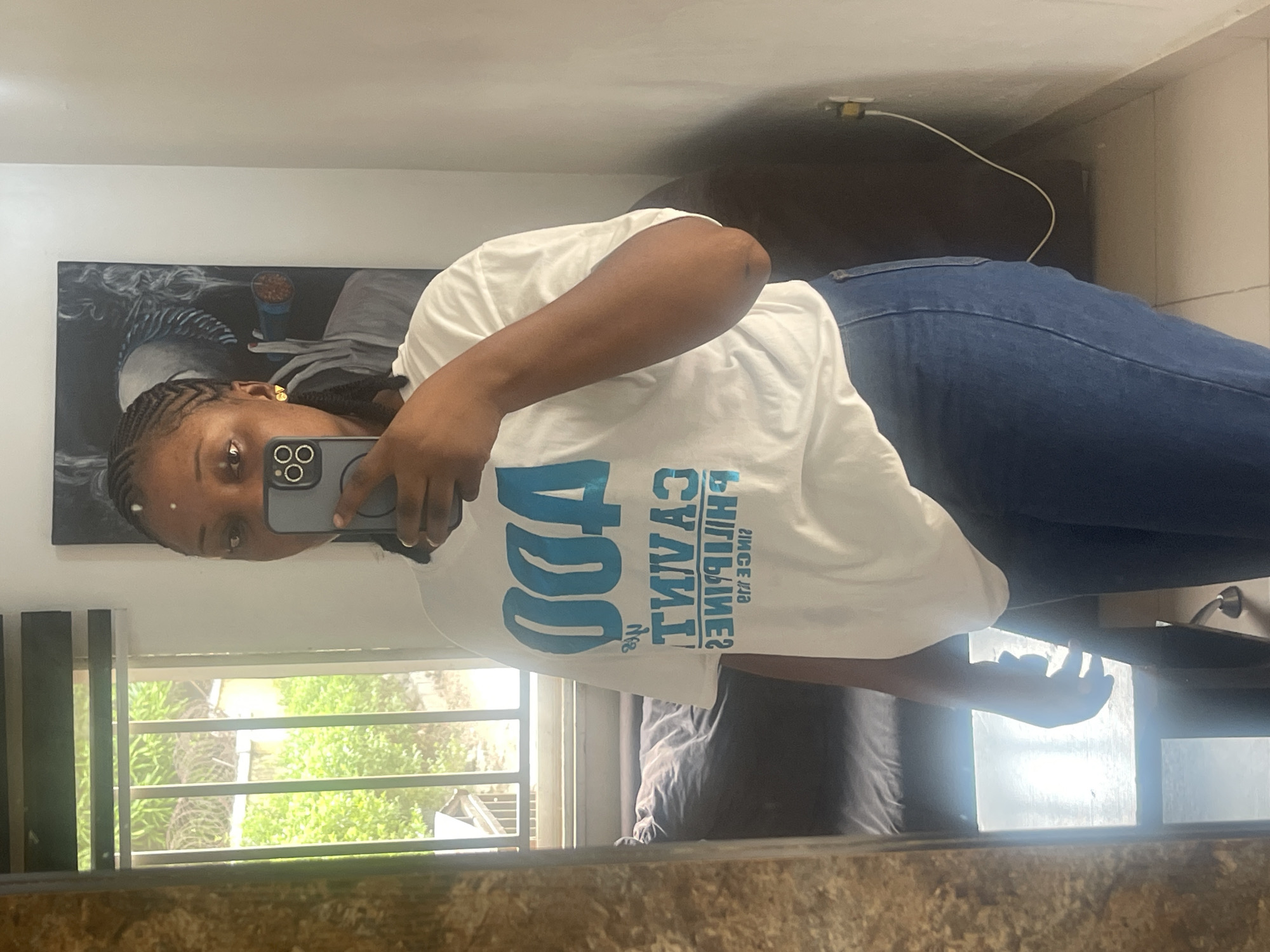A woman is taking a selfie in a mirror. She is wearing a white t-shirt and blue jeans. She has braided hair and is wearing earrings. The reflection shows a window with a view of trees, a painting on the wall, and a sofa. The location appears to be an interior space in Abuja, Nigeria, based on the information provided.