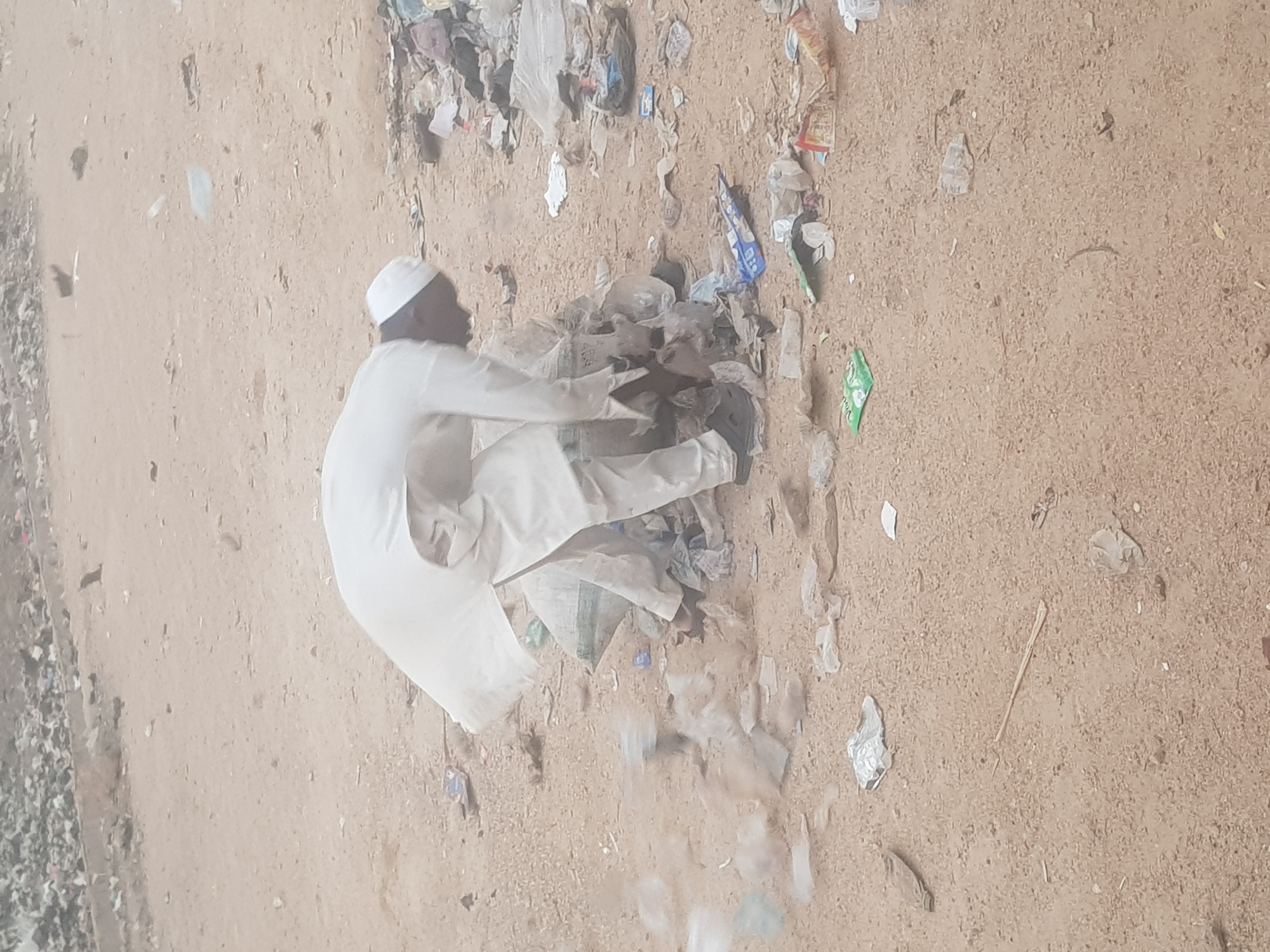 A man wearing a white cap and white clothing is kneeling amidst scattered trash and debris on a dirt or sand surface. The debris includes plastic bags and wrappers. The location is Dutsinma A, Nigeria.