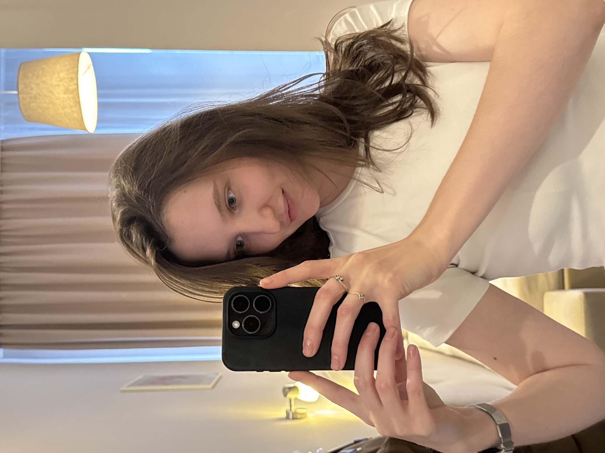 A young woman with brown hair, wearing a white t-shirt, takes a mirror selfie. She is holding a black smartphone with a triple camera system in her right hand. She is wearing two rings on her left hand. The background appears to be a room with soft lighting from a lamp and a window showing a bright, possibly daytime sky. The scene suggests a casual, personal moment. The presence of a lamp and the muted colors of the background elements like curtains and furniture could indicate an indoor setting, possibly a hotel room or a bedroom. The overall mood is relaxed and informal.