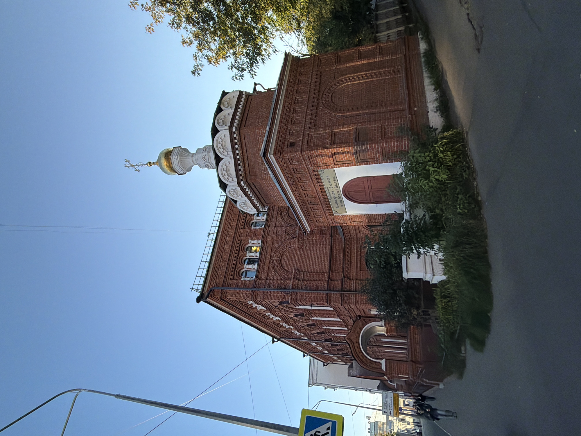 The image shows a brick building with a golden dome and cross, likely a church, in Nizhny Novgorod, Russia. The building is made of red brick and features arched windows and decorative brickwork. A red door is visible on one side of the building. In front of the building, there is a sidewalk and a street. A street sign with pedestrian and traffic symbols is visible on the left side of the image. Several people are walking on the sidewalk. Trees and foliage are present, indicating an outdoor setting. The sky is clear and blue.