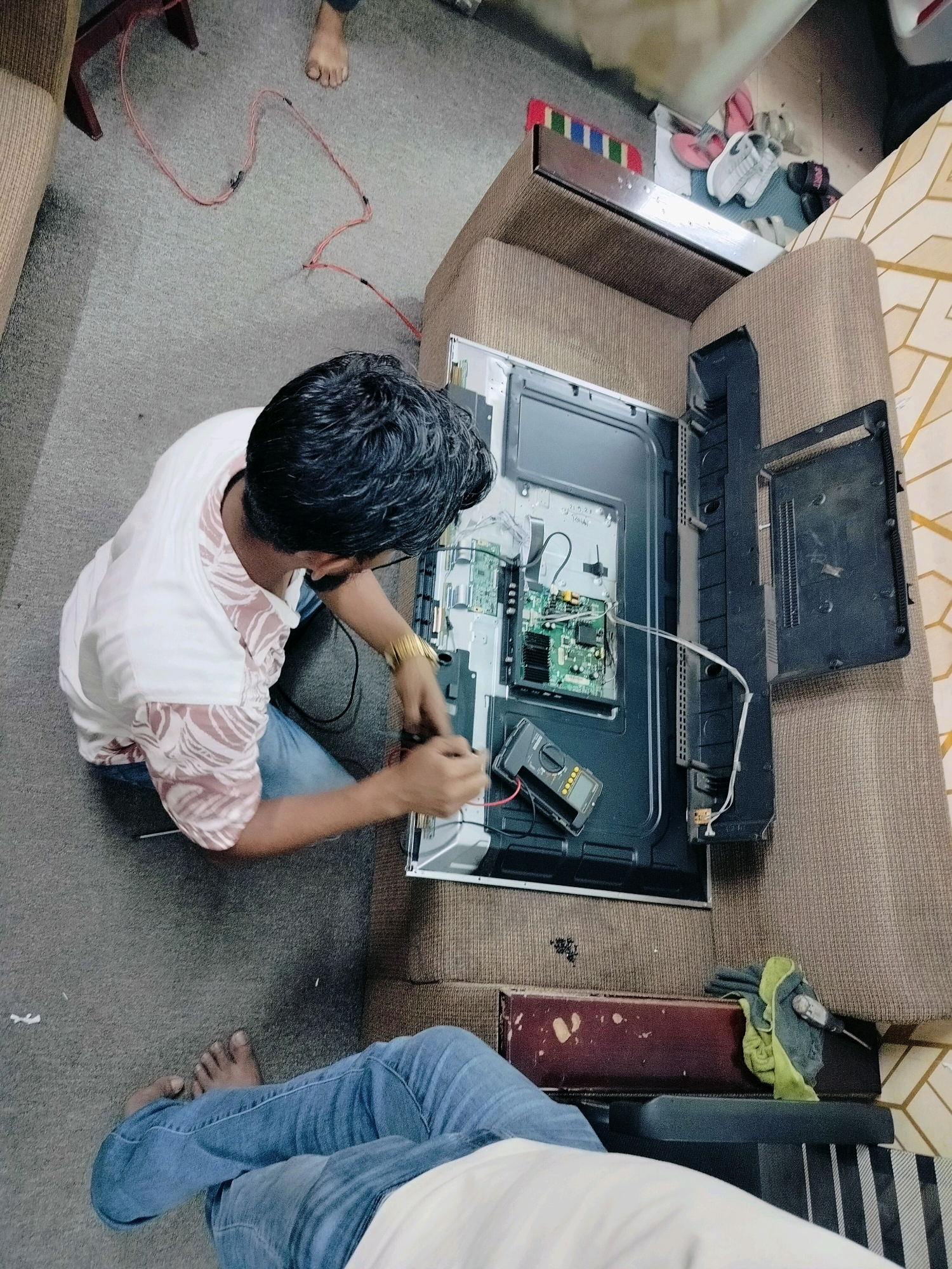 In the city of Dhaka, Bangladesh, an individual is kneeling on the floor and working on an open television set that is lying face up on a couch. The TV's back panel is removed, exposing the internal circuit boards and wiring. A multimeter is placed near the circuit boards, suggesting diagnostic work is underway. The individual is wearing a white t-shirt with a patterned design and blue jeans. Their right hand is engaged with the TV's internal components, while their left hand is resting. In the background, another person's legs, clad in blue jeans, are visible, also seated on the floor. A red electrical cord snakes across the grey carpeted floor. Various items are scattered in the environment, including a red and white striped rug, a pair of flip-flops, and sneakers near the base of a piece of furniture. The scene appears to be an indoor setting, possibly a living room or workshop, with the couch serving as a makeshift workbench.