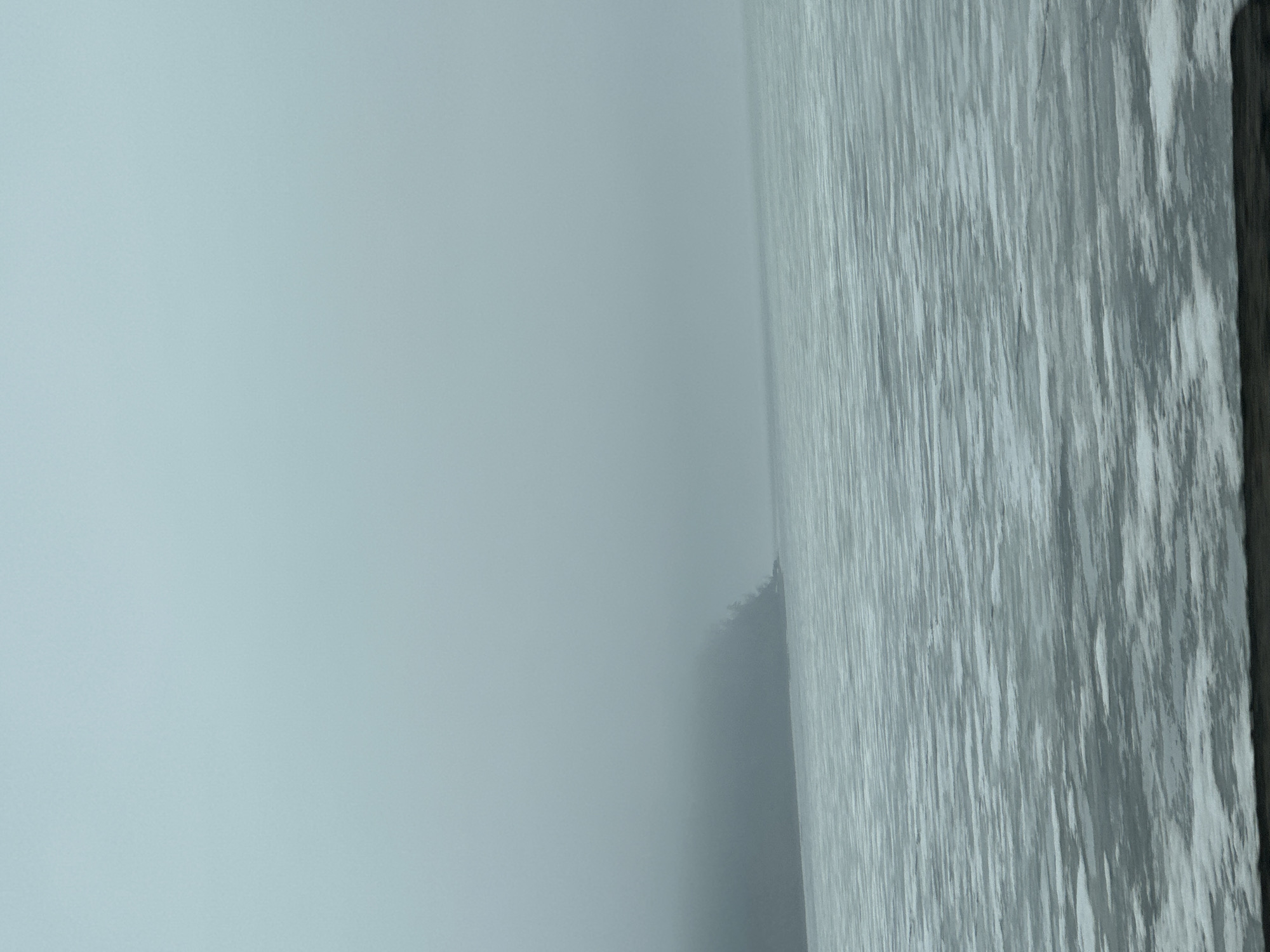 The image shows a body of water, possibly a lake or river, covered in fog or mist. The water's surface is rippled and appears textured due to the light reflecting off the water. In the background, a hazy landmass is faintly visible. The scene is muted in tone, with shades of gray and blue dominating. The location is assumed to be Chaikovsky, Russia.