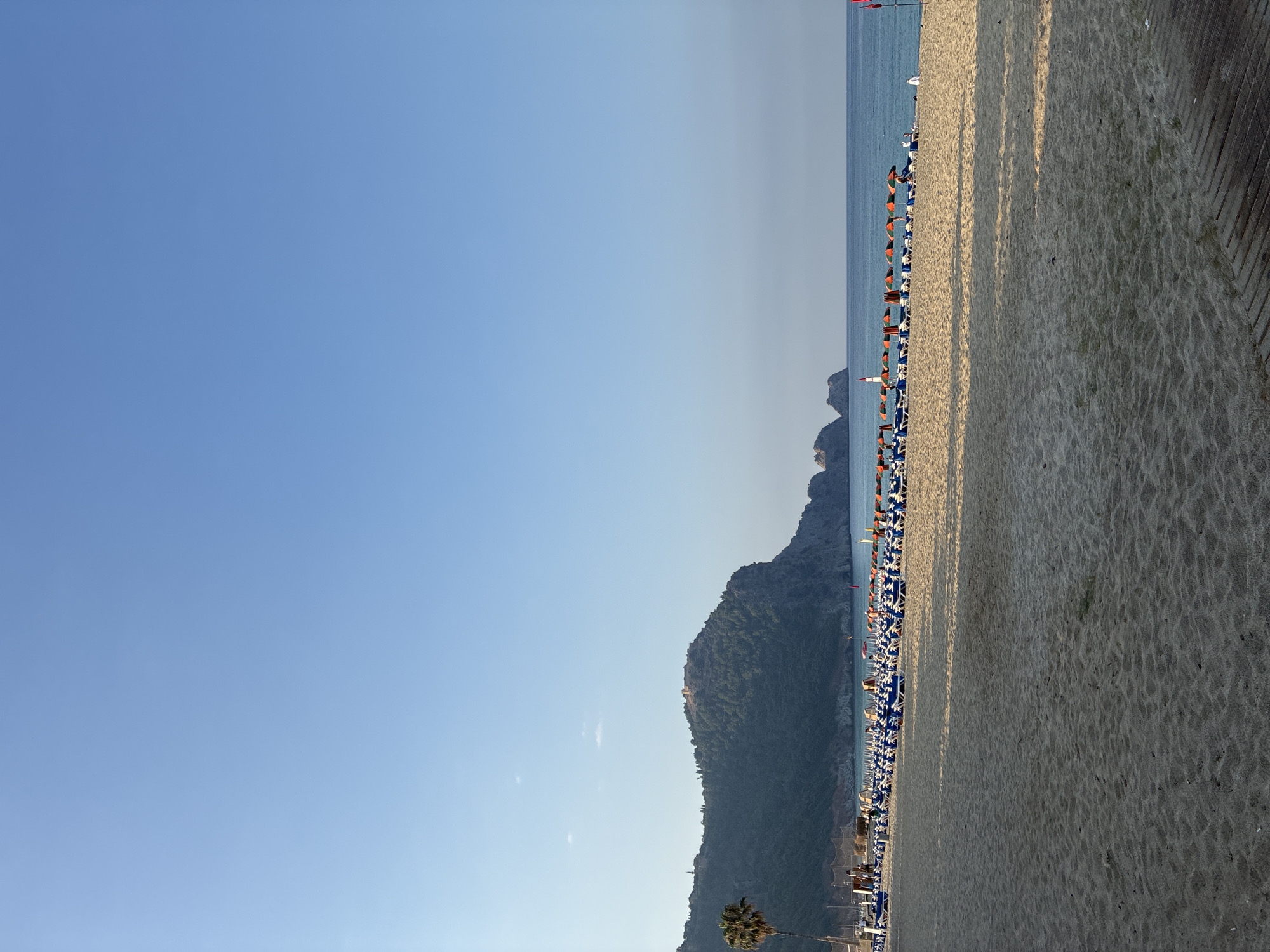 The image shows Cleopatra Beach in Alanya, Turkey, under a clear blue sky. The sandy beach is lined with numerous blue and white lounge chairs and colorful umbrellas. In the background, the sea is calm and a large, tree-covered hill with ancient ruins on its summit dominates the horizon, characteristic of Alanya Castle. A single palm tree stands on the left side of the beach. Several people can be seen scattered among the chairs and walking on the beach. The lighting suggests it is late afternoon, with long shadows cast across the sand. A wooden walkway is visible on the right side of the foreground.