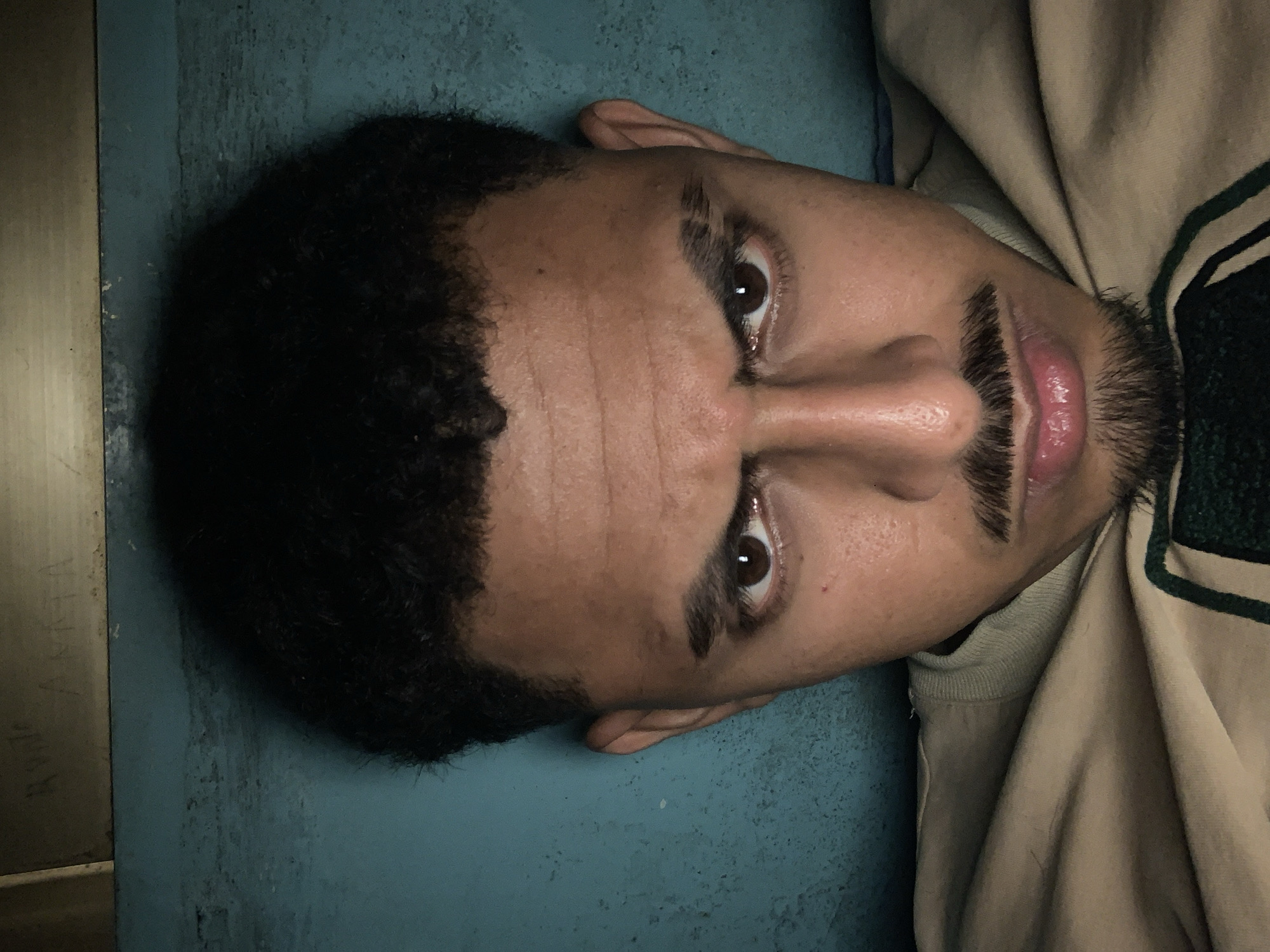 A rotated close-up portrait features a young adult male looking directly into the camera. He has dark, curly hair, dark brown eyes, and prominent eyebrows. His face is clean-shaven except for a well-groomed mustache and a small chin beard/goatee. His lips are slightly parted, revealing a pink hue. He appears to have a neutral or slightly serious expression.
The subject is positioned with his head resting against a textured, teal-blue surface, which appears to be a wall or a large panel. To the left, there is a lighter, vertical panel, possibly made of wood or metal, which shows signs of wear and faint, illegible markings or scratches. One crop reveals faint text on this surface, which is difficult to discern but might contain letters like "RVM" and "ALTA."
He is wearing a beige or light brown sweatshirt or hoodie with a green stripe visible on the right side, near his shoulder. The lighting is soft and somewhat dim, suggesting an indoor setting or low-light conditions. The overall composition is a direct, head-and-shoulders shot. The scene is set in Santa Ana, Venezuela.