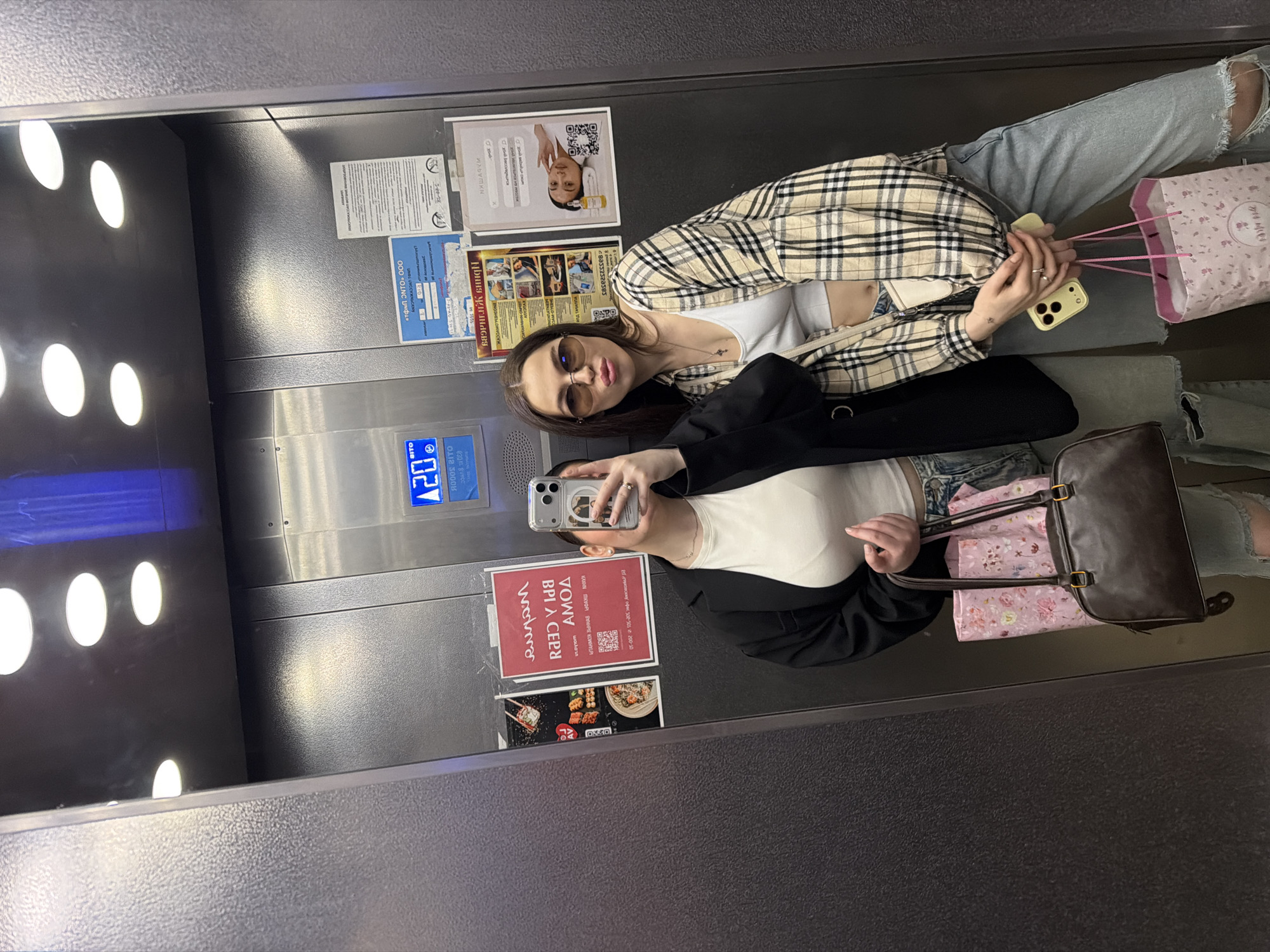 The image depicts two young women taking selfies in an elevator. The elevator has stainless steel walls with various notices posted on them. One woman is wearing a white top, a black blazer, and jeans, holding a brown handbag and a pink shopping bag. The other woman is wearing a white top, a plaid jacket, and ripped jeans, also holding a pink shopping bag. A sign on the elevator wall includes Russian text and the address "Amosova street, Chaikovsky". The elevator has round ceiling lights and a digital display indicating the floor number.