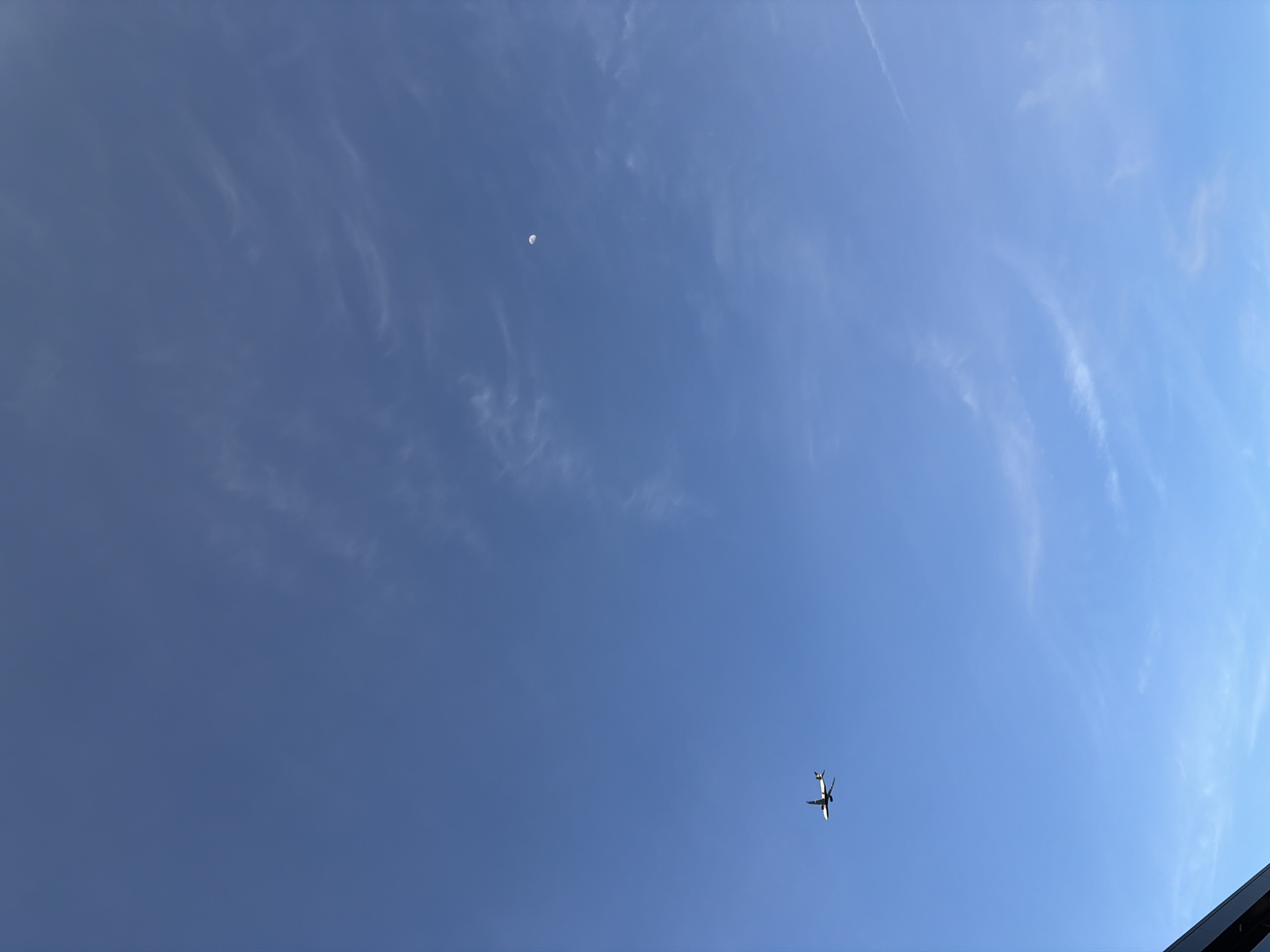 A low-angle, wide shot depicts a bright, clear blue sky with wispy cirrus clouds sparsely scattered. A small crescent moon is visible in the upper left quadrant. An airplane, likely a passenger jet, is positioned in the lower right quadrant, appearing to be in flight. No people are visible. There are no other discernible objects or actions in the scene. The specific location within the city of Montreal, Canada cannot be determined from the image.