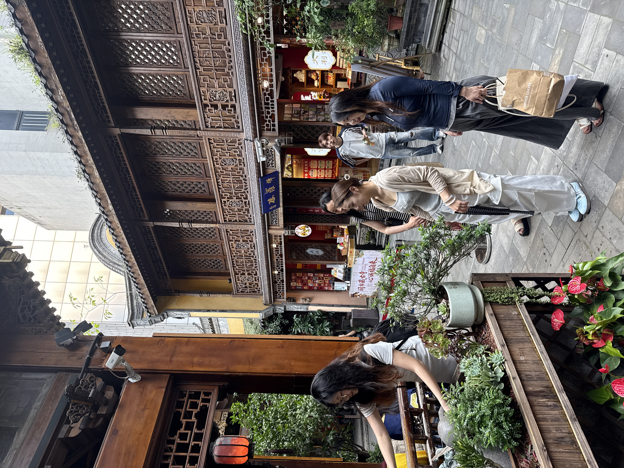 The image captures a vibrant street scene in what appears to be a historic commercial district, likely in Yuzhong District, China. The setting is dominated by multi-story traditional Chinese wooden buildings, characterized by their richly carved facades, intricate lattice patterns, and overhanging roofs with decorative eaves. These structures line a paved alleyway, creating a dense, enclosed atmosphere. In contrast to the traditional architecture, a modern, light-colored building is visible in the upper left background, and security cameras are mounted on wooden beams, blending old and new urban elements.
The scene takes place during the daytime, under what seems to be overcast or shaded conditions, resulting in soft, diffused light. Several people are present, engaging in casual activities. In the foreground, a woman with long dark hair is bending over a wooden planter filled with a variety of plants, including succulents and red anthurium flowers, suggesting she might be tending to them or admiring them. Further down the alley, three individuals are seen. One woman wears a dark blue long-sleeved top and a long grey skirt, carrying a brown paper bag with "MANNER COFFEE" printed on it, along with other shopping. Another woman is dressed in a light-colored cardigan over a grey top and white pants, wearing light blue platform sandals. A third person, seen further back near a shop, wears a white t-shirt with black striped sleeves and jeans.
The shops along the street display merchandise through glass fronts and feature various signs. One prominent blue and gold sign above a shop entrance contains Chinese characters that appear to read "聚德昌" (Jùdéchāng). Another shop displays a single Chinese character "利" (Lì). Additional smaller signs with red backgrounds and white Chinese text, as well as promotional posters, are also visible on the shopfronts. The ground is paved with grey stone tiles. The overall impression is one of a lively, culturally rich commercial area where people are browsing or passing through.