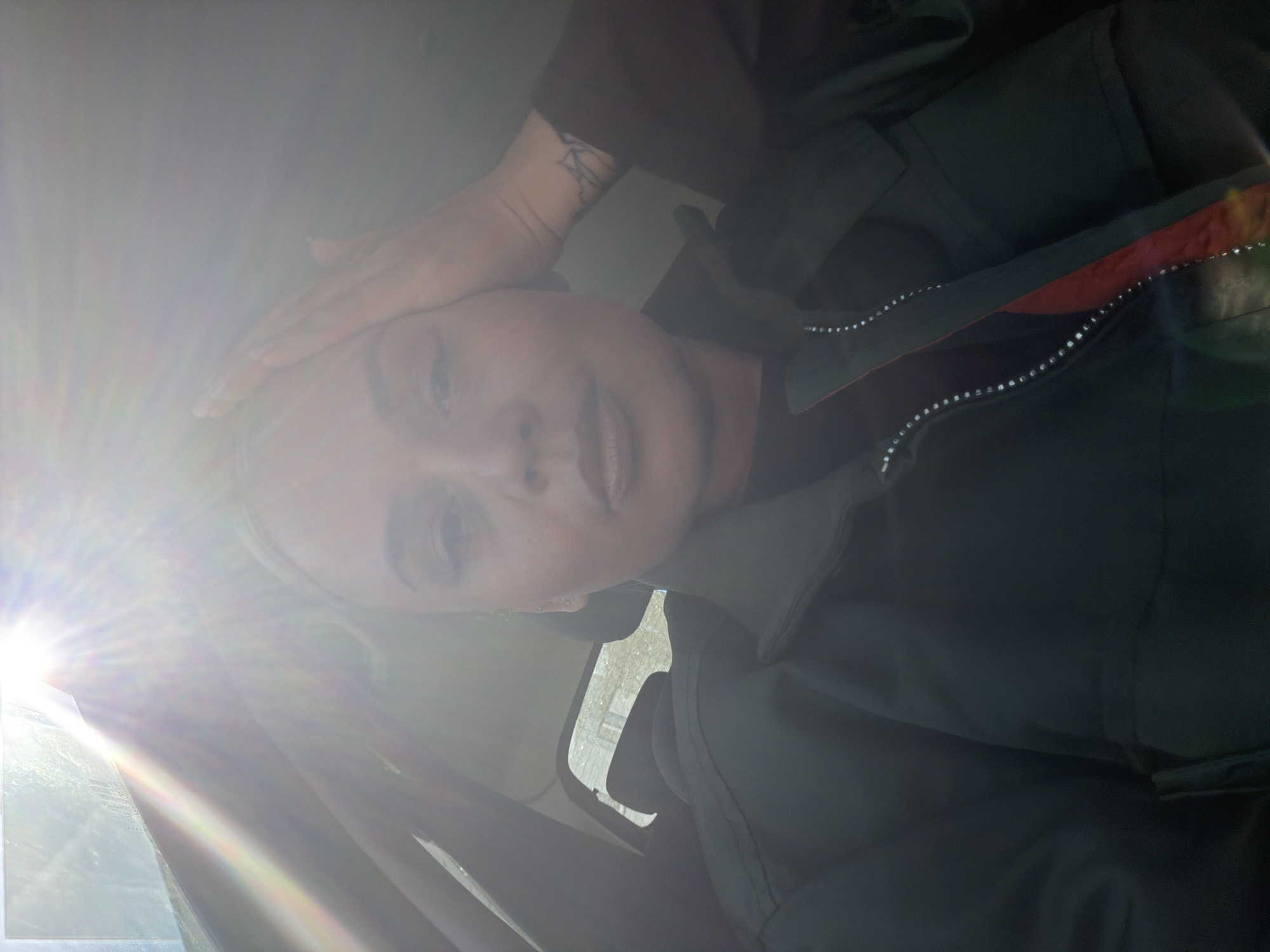 A fair-skinned person with blonde hair is sitting inside a vehicle, possibly a car, and obscuring her face with one hand to block out the strong sunlight. The person is wearing a dark green zip-up jacket, visible in the foreground. A portion of a wrist tattoo can be seen on her left wrist. Outside the partially-opened vehicle window, a building and foliage can be vaguely seen. Without further information, it's impossible to confirm if the location is Sarapul, Russia.