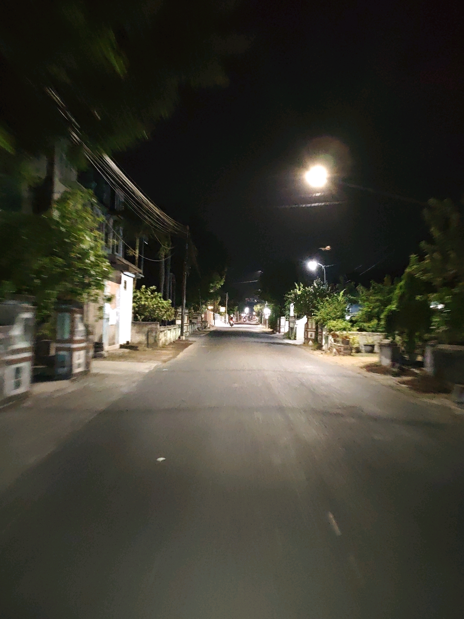 The image depicts a street scene at night in Pagu, Indonesia. A paved road stretches into the distance, flanked by buildings and trees. Streetlights illuminate the scene, casting a warm glow on the asphalt. The motion blur suggests the photo was taken from a moving vehicle. On the left side of the road, a building with multiple windows and a balcony is visible. On the right, there are more buildings and lush vegetation. In the distance, a motorcycle with its headlights on is approaching, and further down the street, several other lights suggest more vehicles or buildings. The overall impression is of a quiet, dimly lit street at night in a residential area.